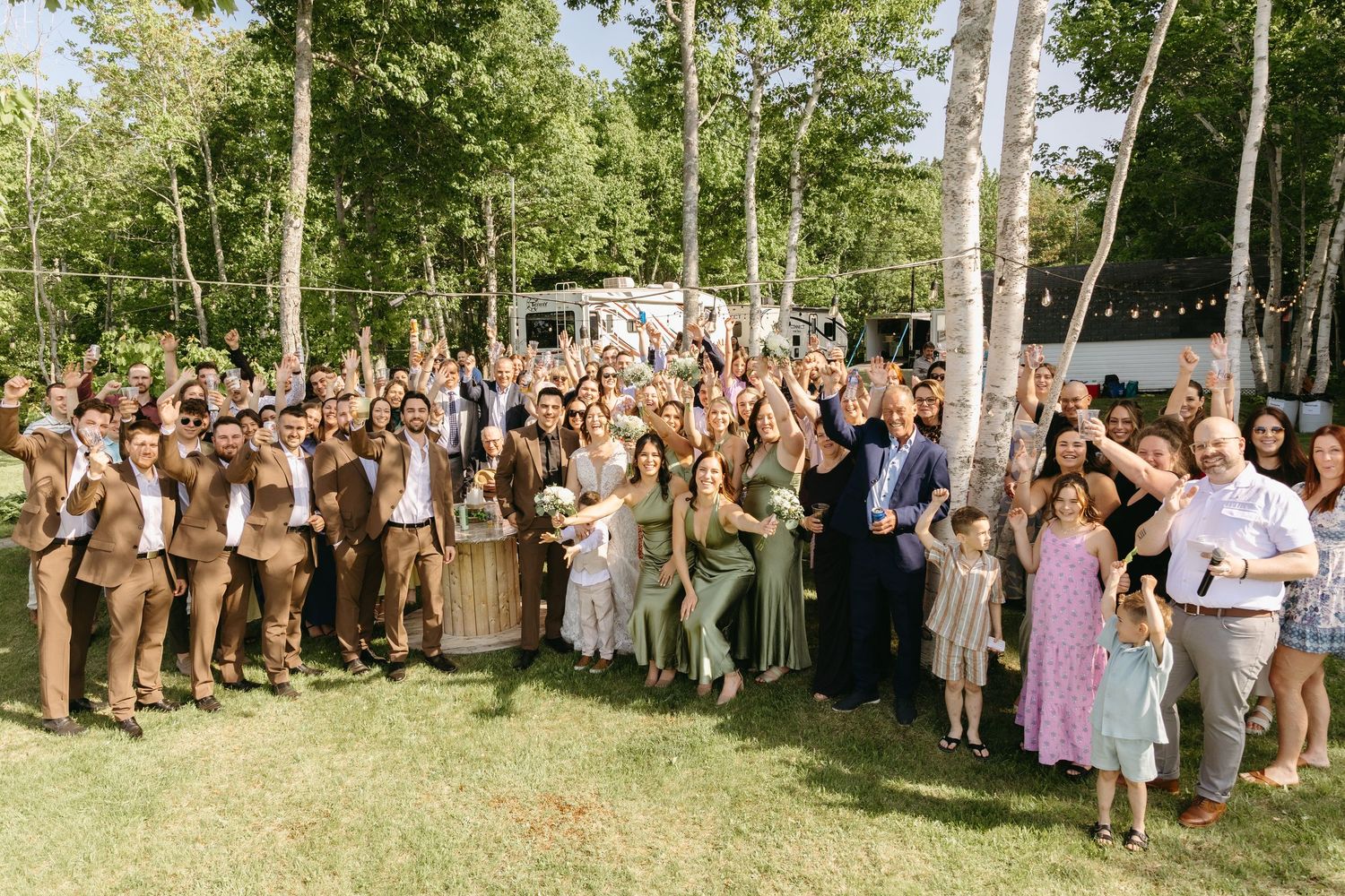 Large group of wedding guests gathered outdoors for a photo beneath tall trees on a sunny summer day.