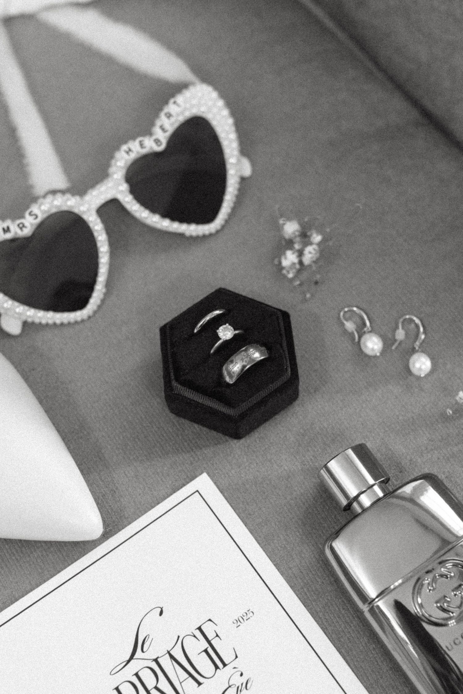 Black and white wedding detail photo showing rings in boxes, sunglasses, and invitation cards on a light surface.