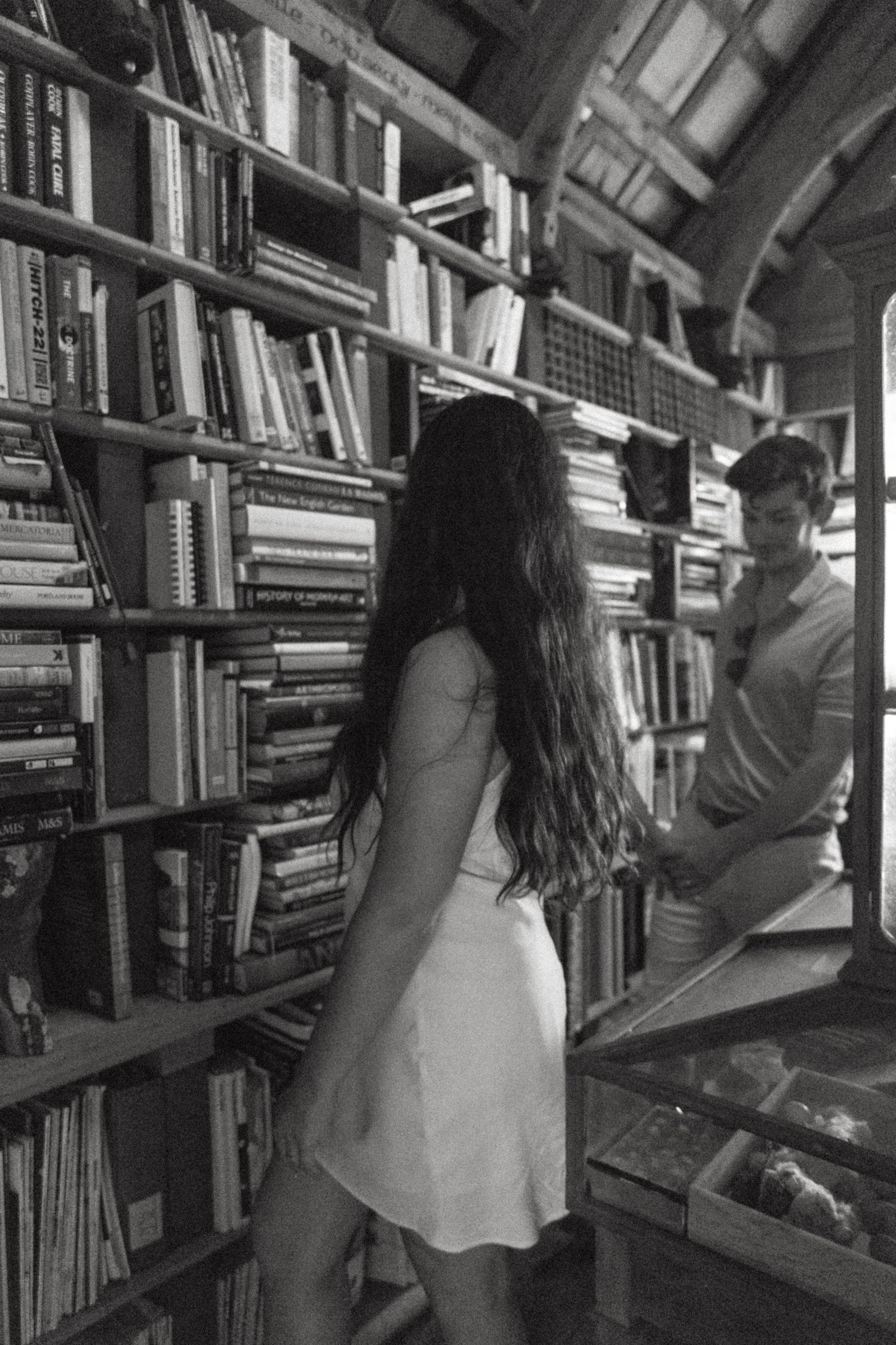 Black and white image of bookshelves in a library with someone browsing through books.