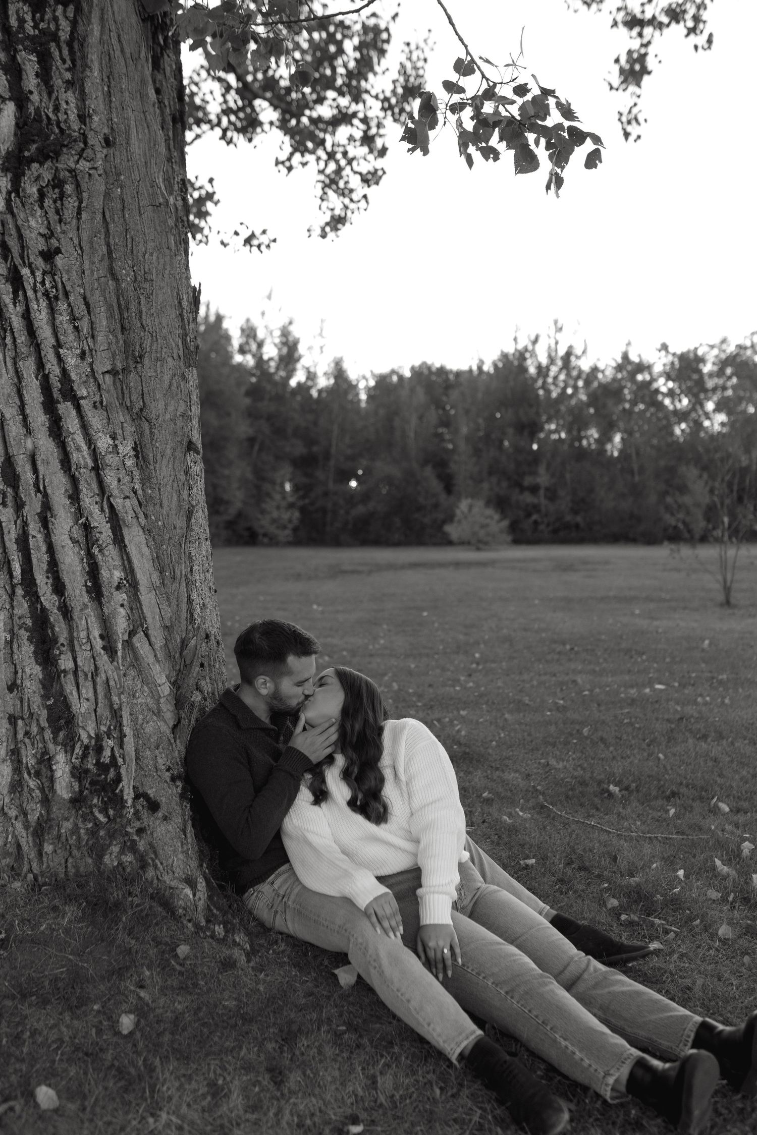 A couple in casual attire shares an intimate moment while resting against a tree trunk in a peaceful park setting.