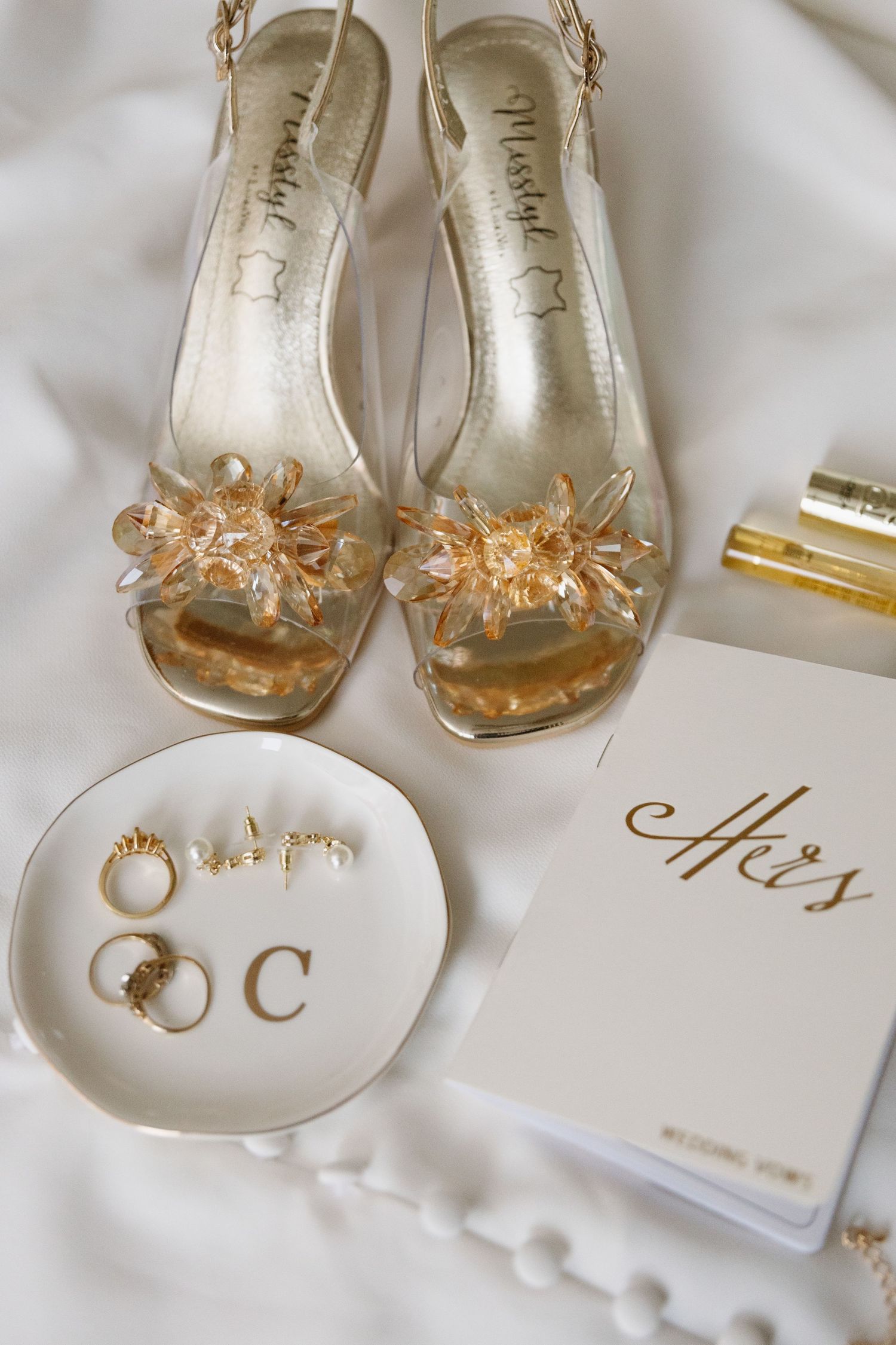Elegant bridal accessories including crystal-embellished peep-toe shoes, wedding rings on a dish, and monogrammed stationery.