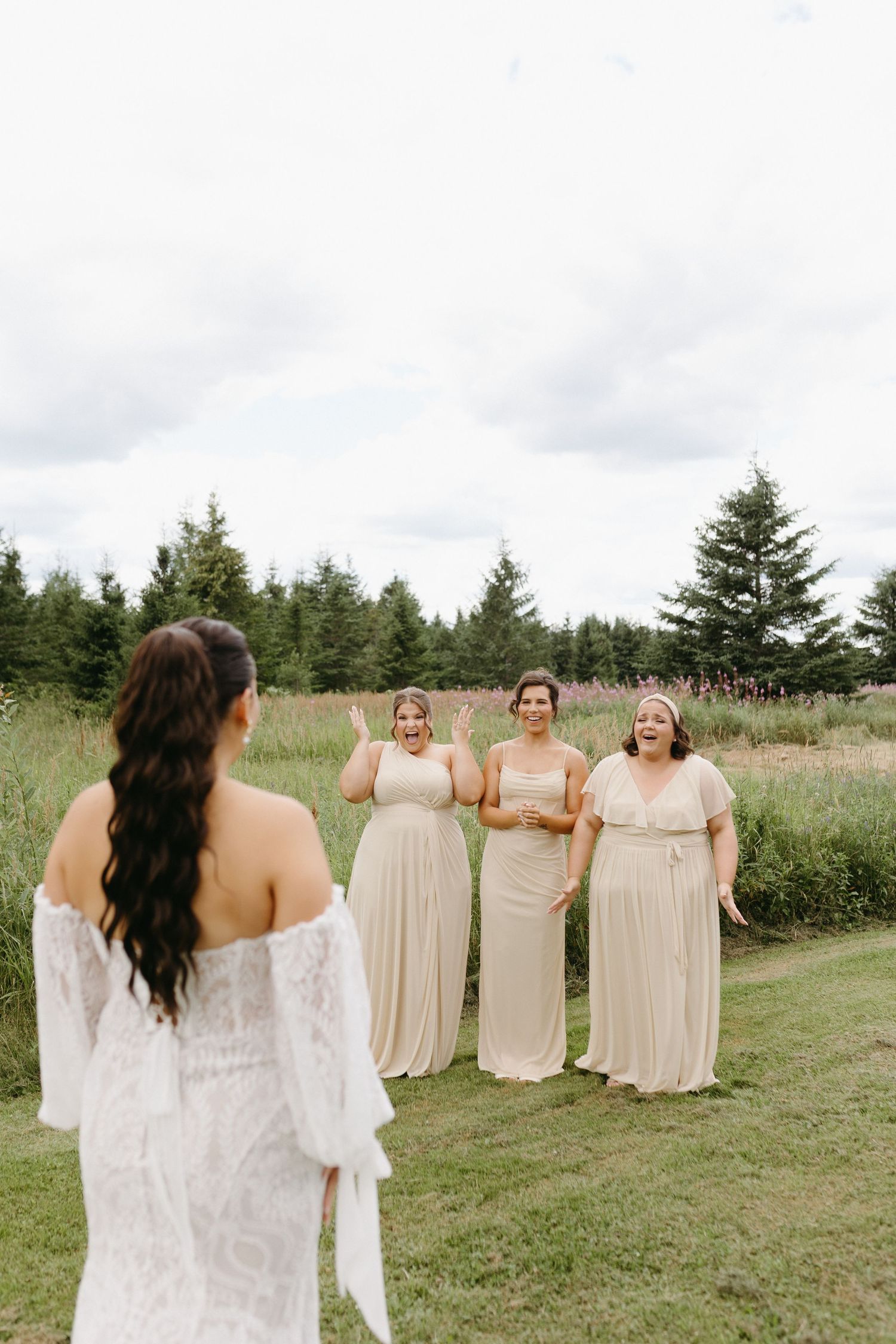 Bridesmaids in beige dresses stand on grassy field watching bride in white lace gown during outdoor wedding celebration.