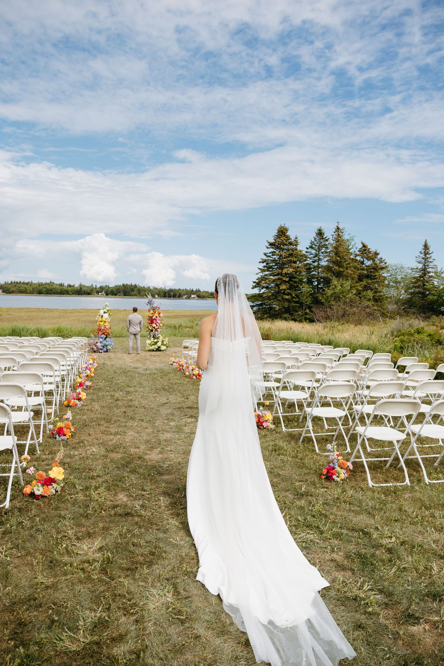 An outdoor wedding ceremony setup with white chairs and floral aisle decorations against a waterfront and evergreen tree backdrop.
