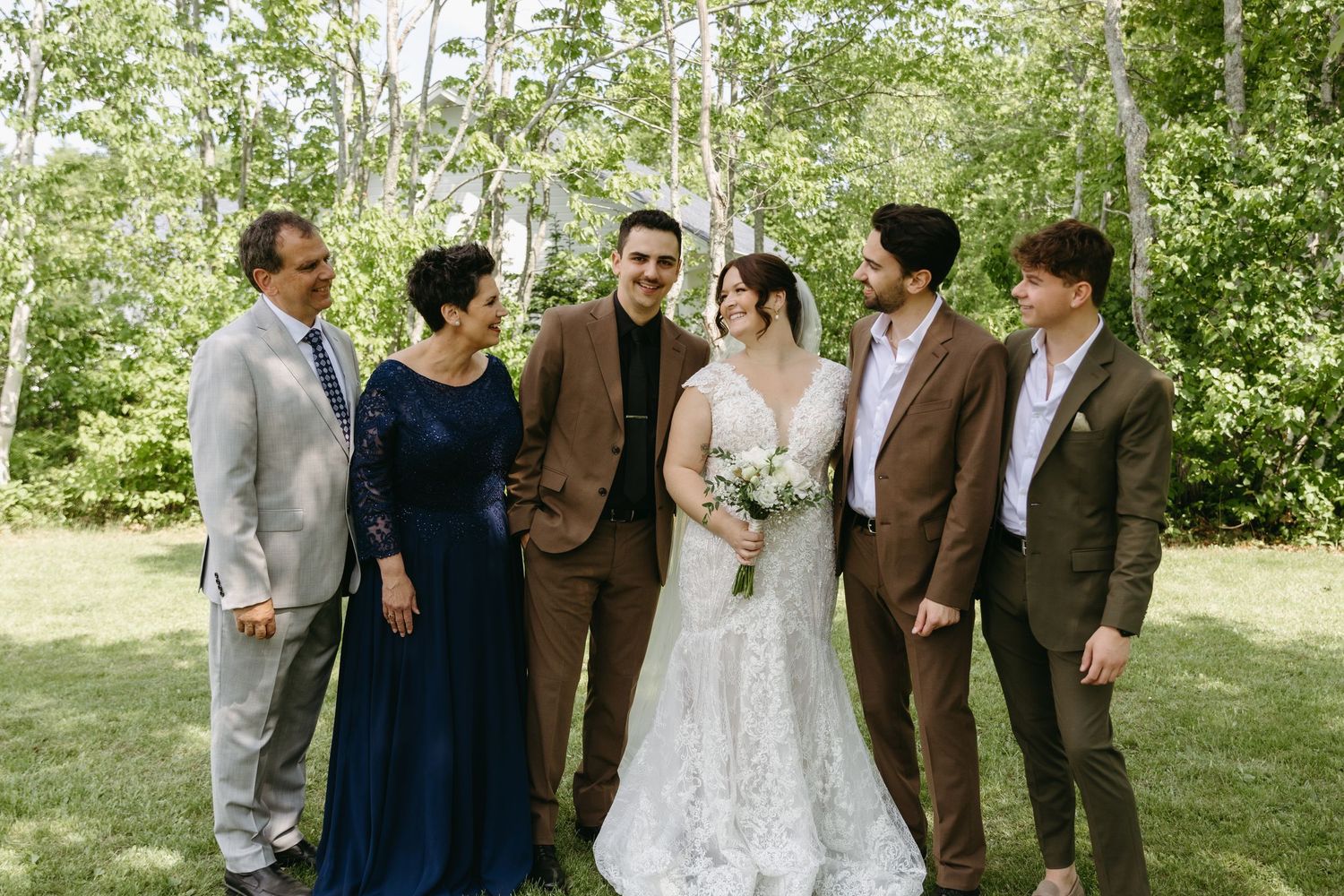 A wedding party stands together outdoors in a garden setting wearing formal attire and brown suits.
