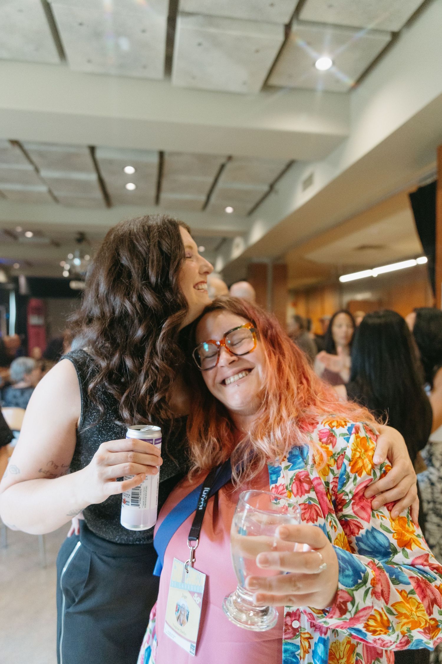 Two friends share a joyful moment at an indoor event, one wearing glasses and a colorful floral outfit while holding drinks.