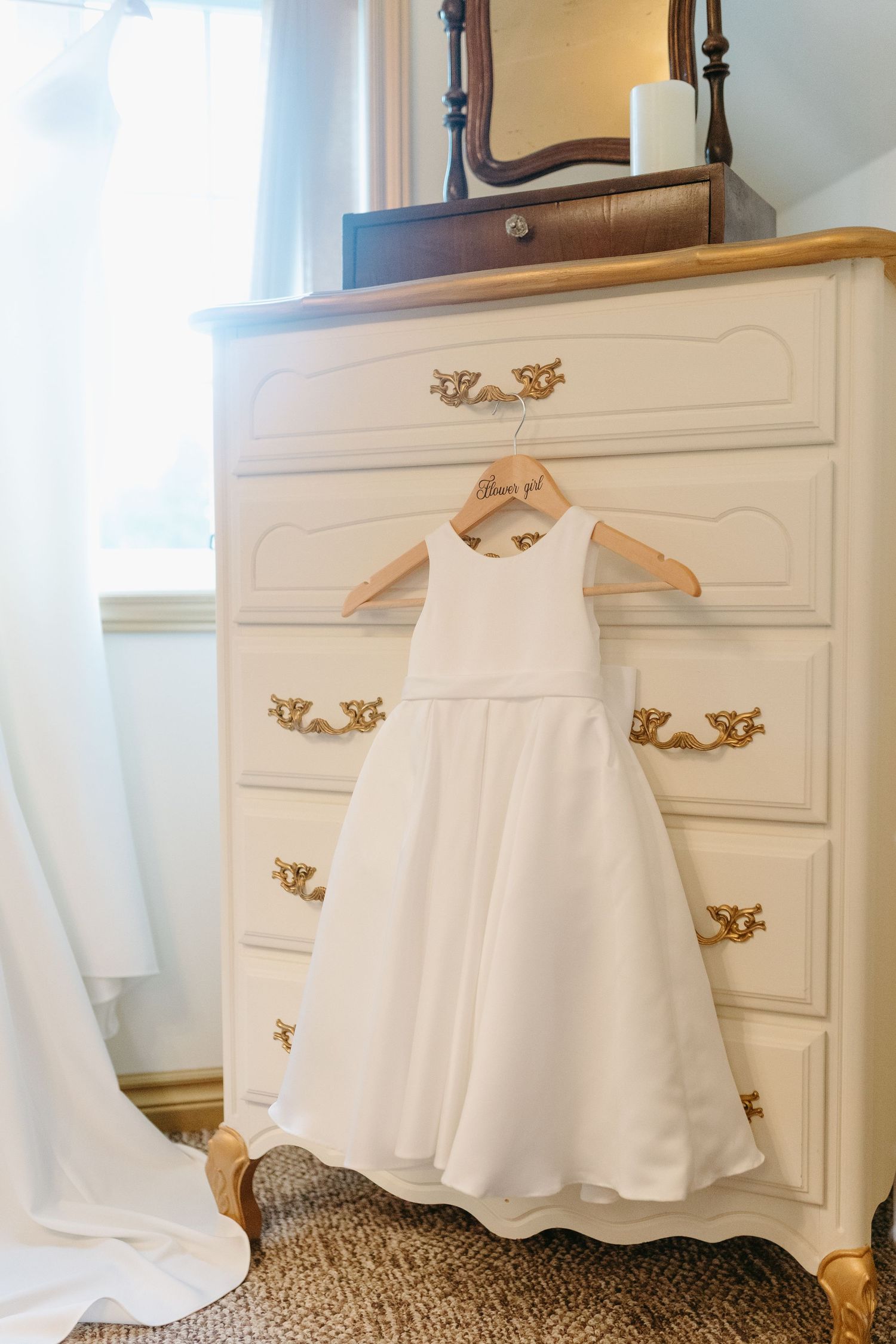 A white wedding dress hangs on display against an elegant white dresser with ornate brass hardware.