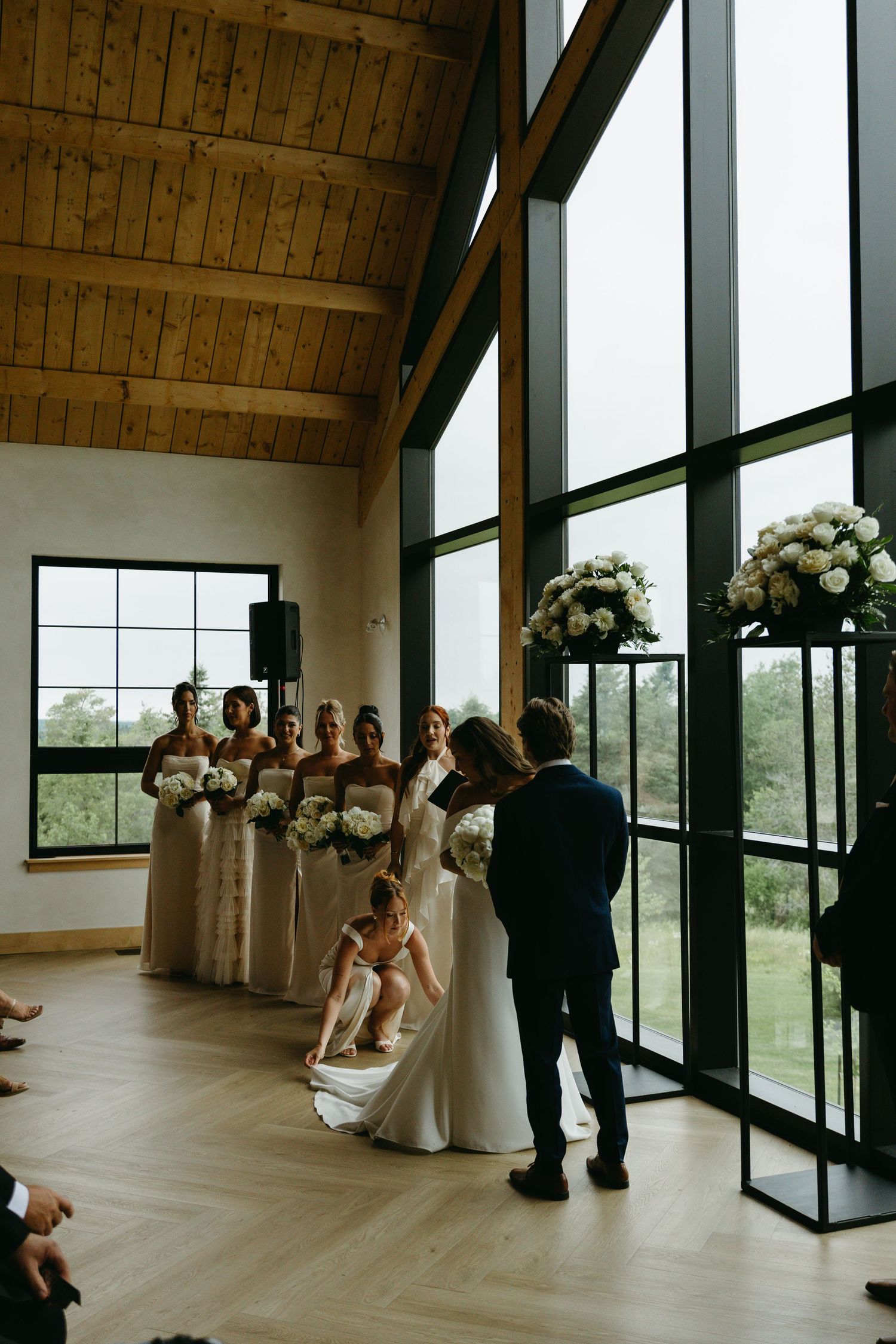 A wedding ceremony takes place in a modern venue with floor-to-ceiling windows, wooden beams, and elegant floral arrangements.