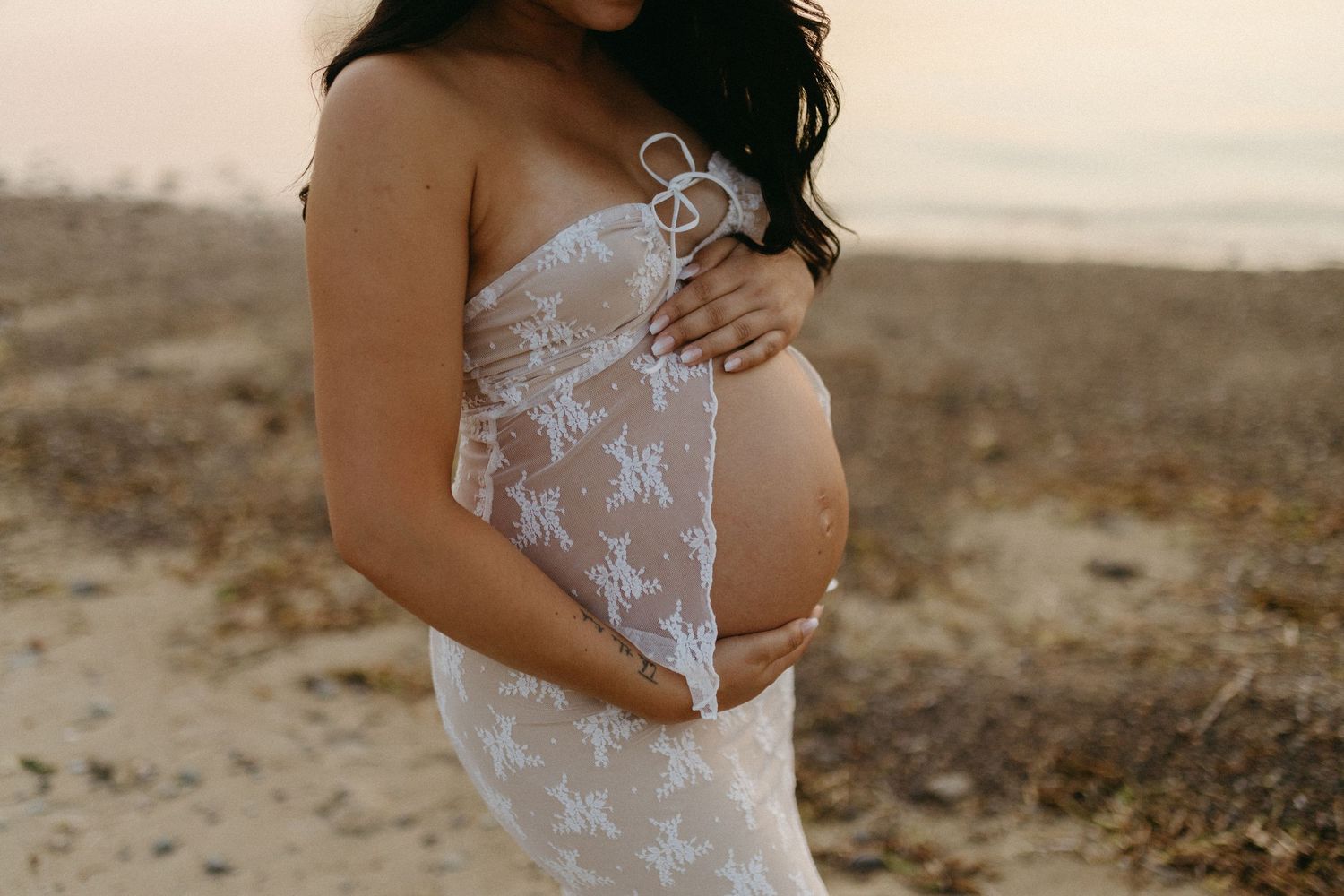 Expectant mother in white lace dress poses on beach during sunset maternity photoshoot.