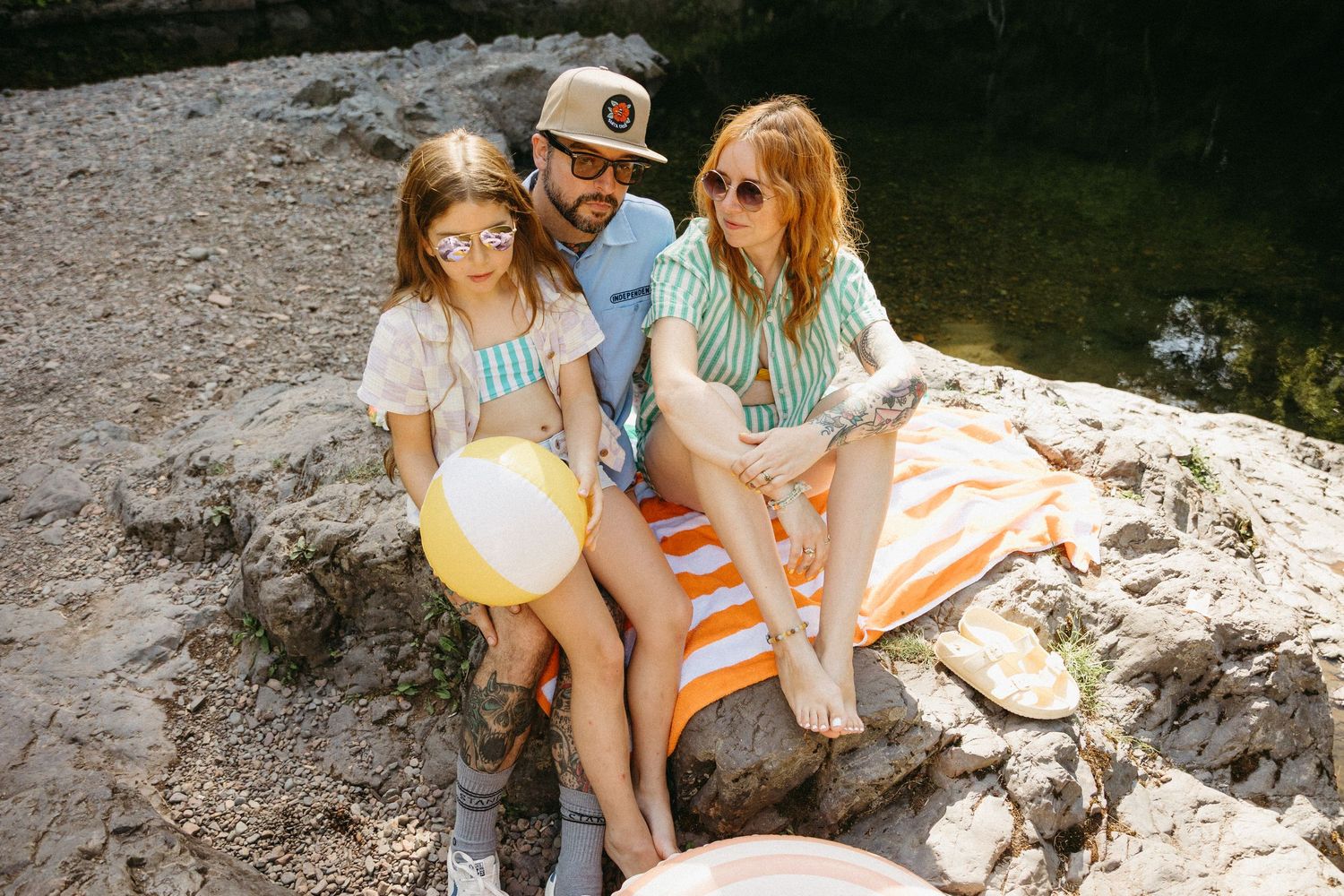 Group enjoying a sunny day by the water sitting on rocks with a yellow beach ball and orange striped towel.