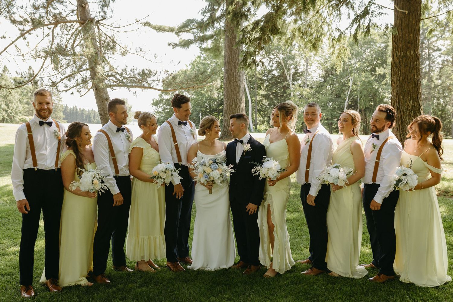 Wedding party poses outdoors under tall trees wearing white dresses and suits with suspenders.