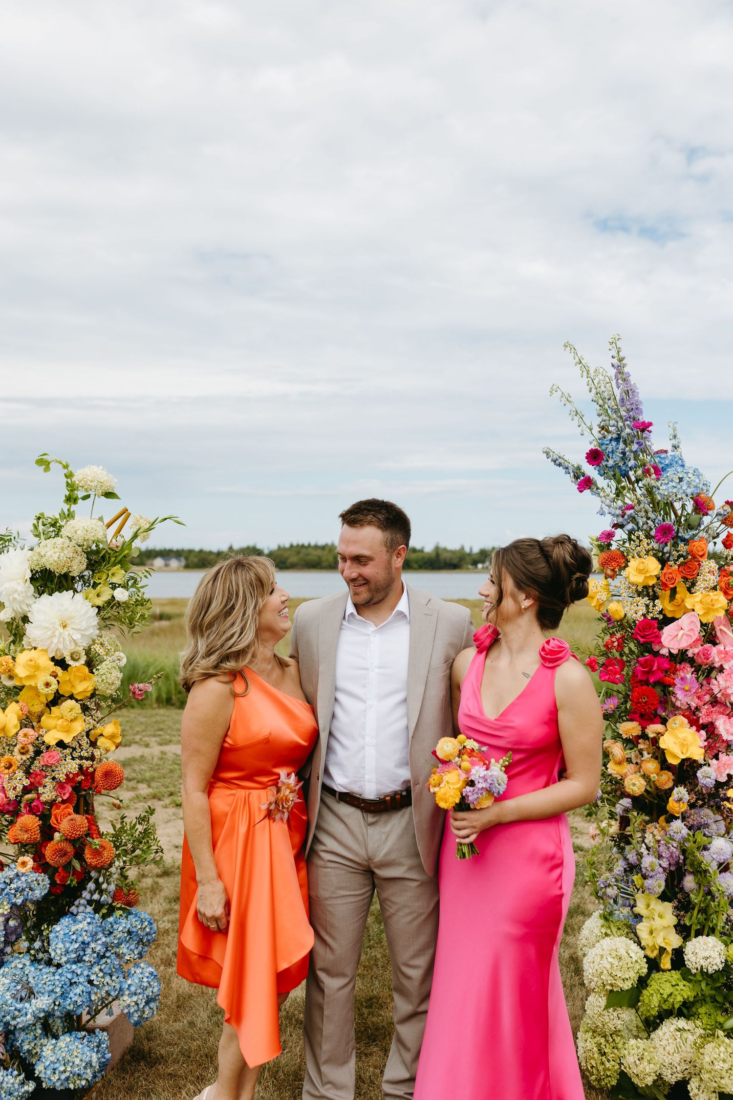 Wedding party poses together outdoors with vibrant orange, pink and yellow floral arrangements against cloudy sky.
