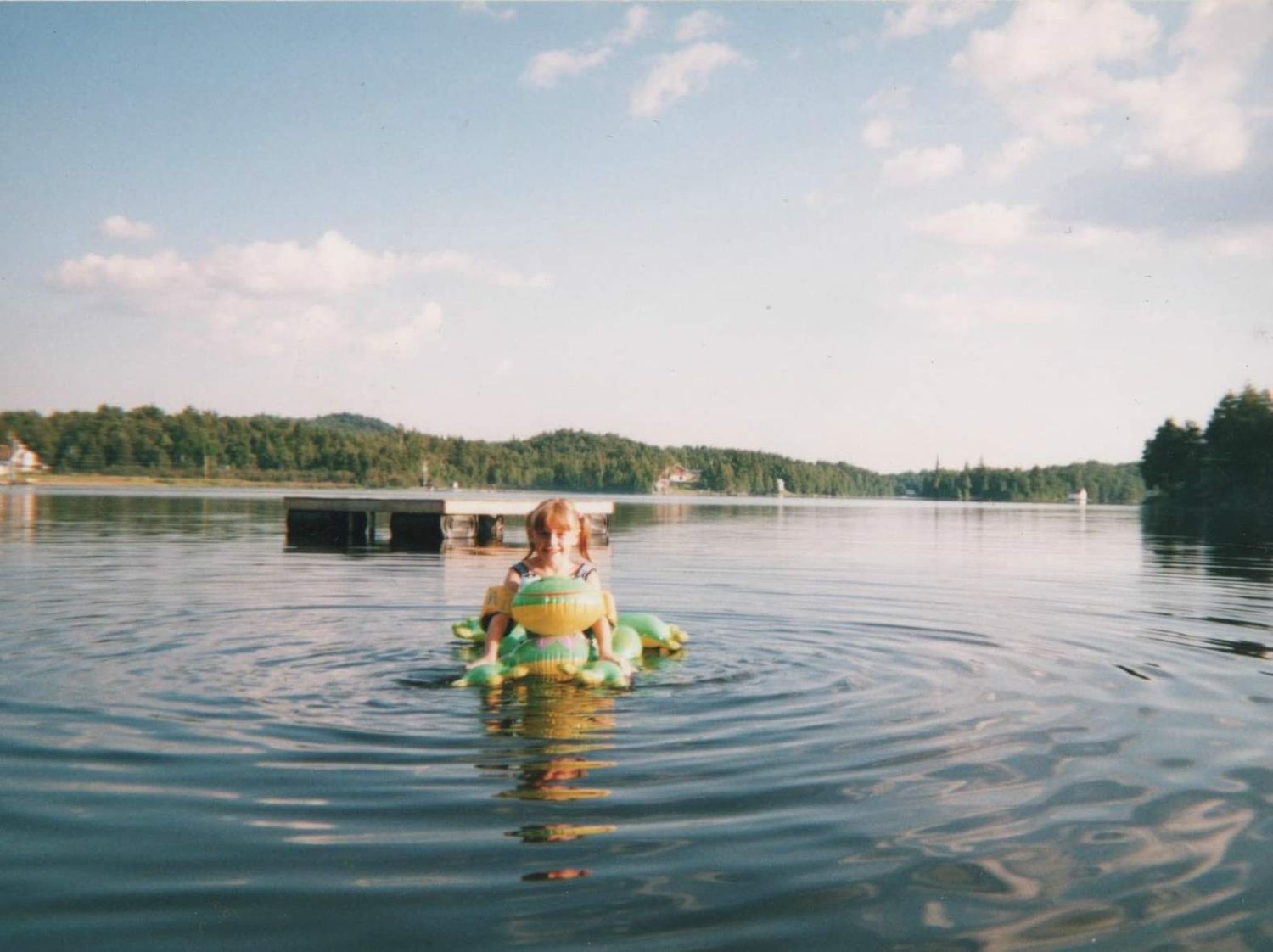 Panoramic view of people tubing on a calm lake with evergreen trees lining the shoreline.