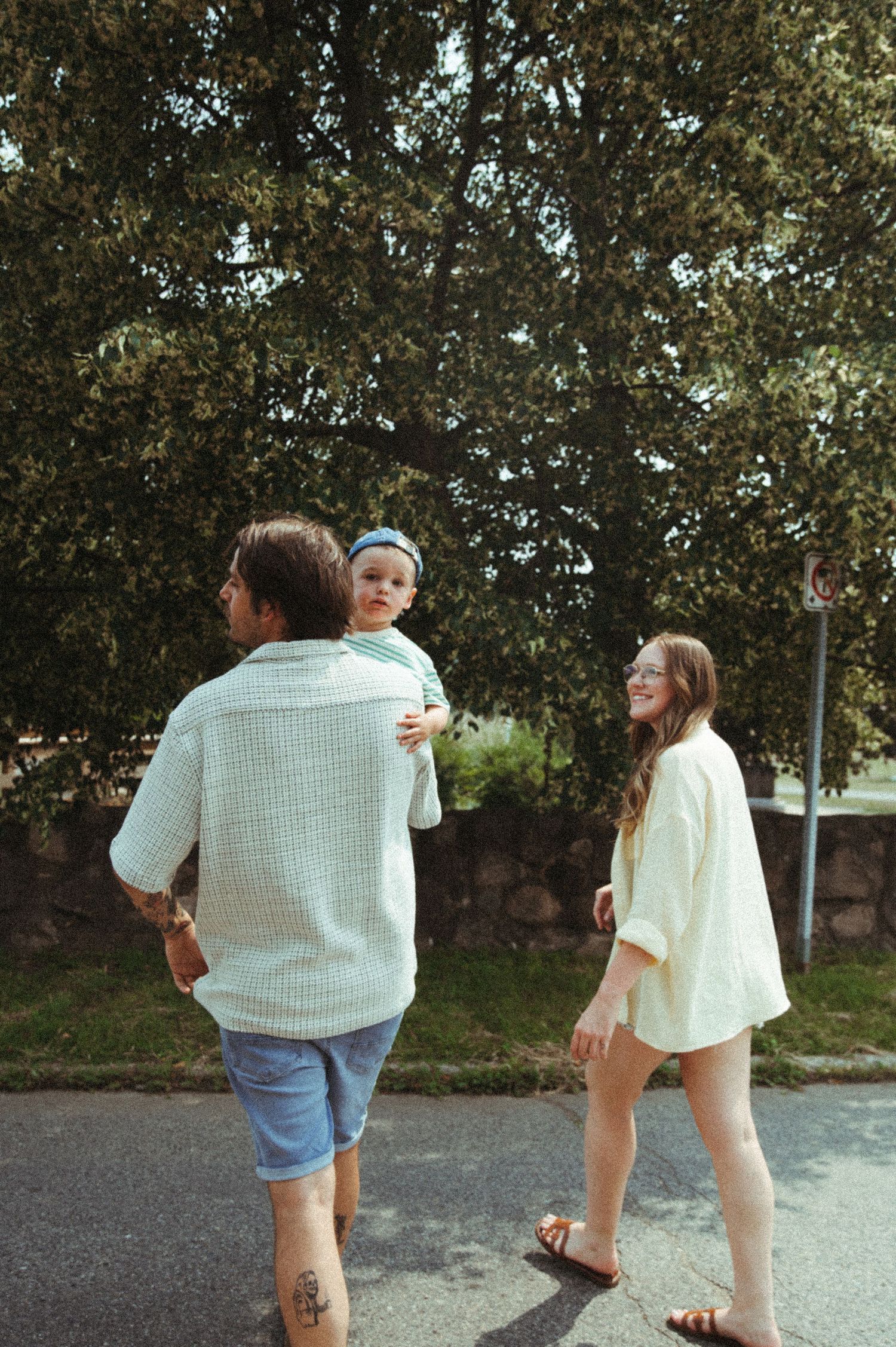 A family walks together on a sunny day along a tree-lined street, wearing casual summer clothing.