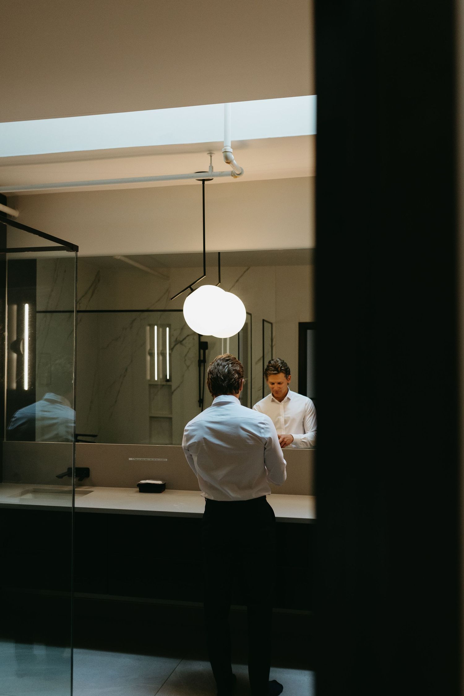 A person looks in a bathroom mirror with a round pendant light hanging above, creating a moody interior reflection.