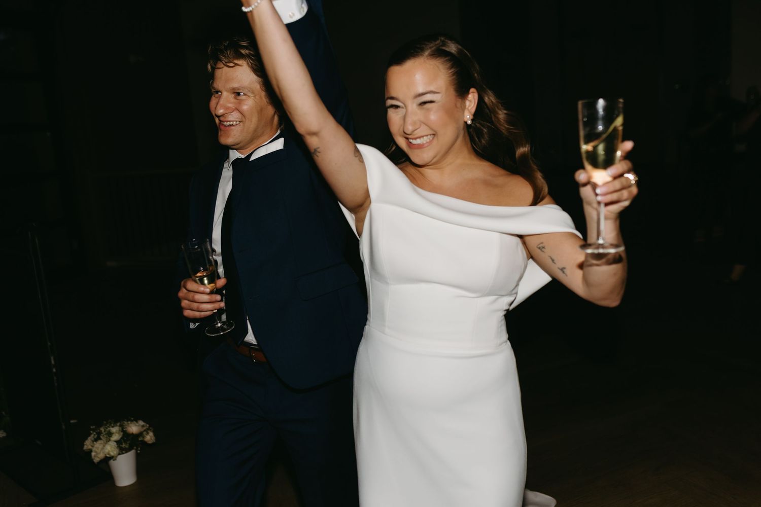 A couple celebrates at a wedding reception, with raised champagne glasses and joyful expressions in the dark.