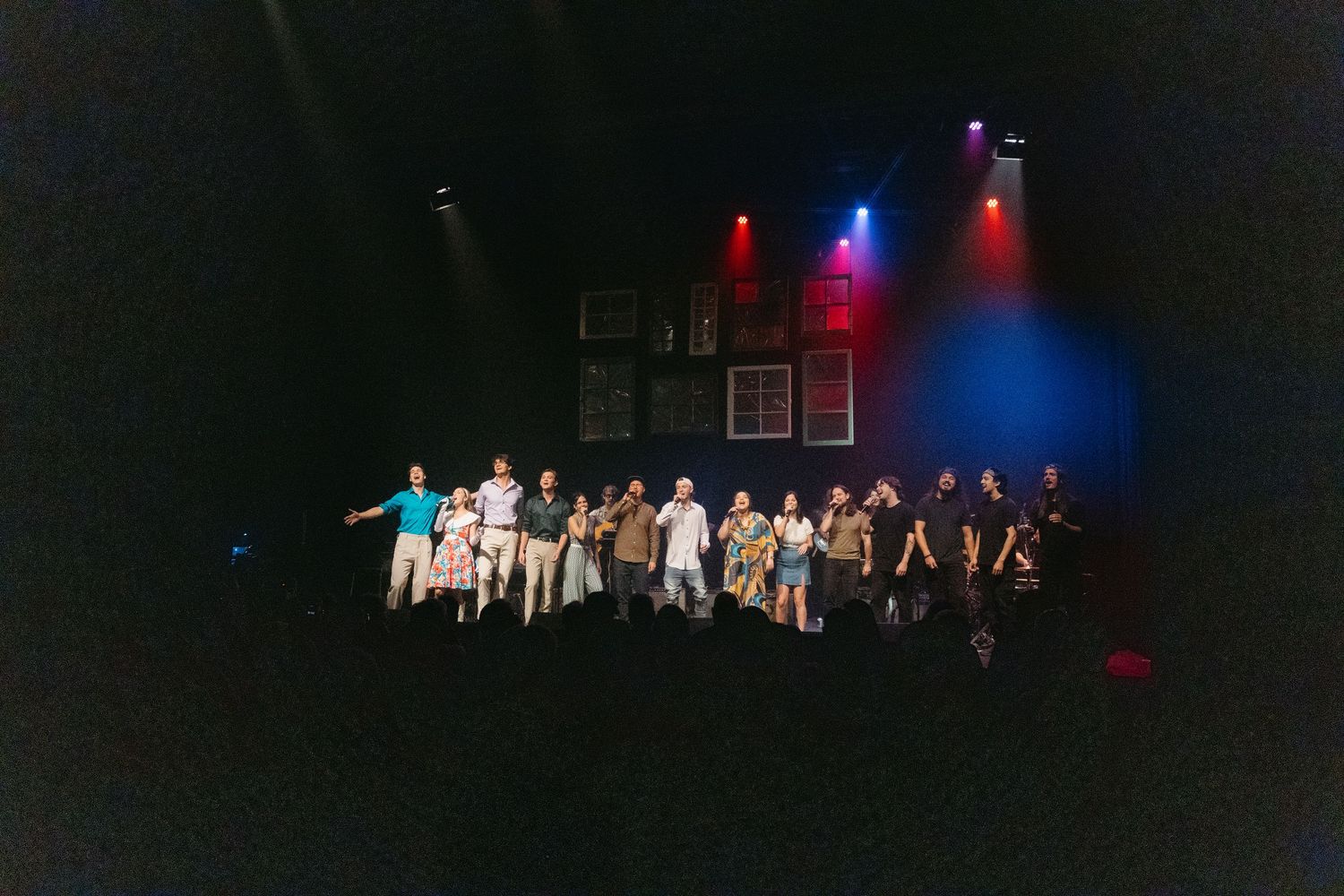 Performers in white outfits stand on a dark stage illuminated by blue and red theatrical lighting during a show.