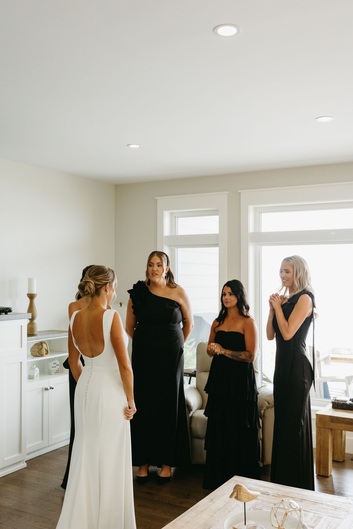 Wedding party members in black dresses gather around a bride in white in a bright modern room.