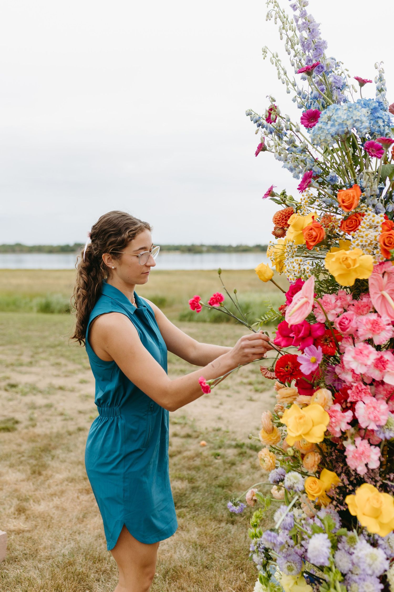 A floral designer in a teal dress arranges a vibrant outdoor installation with pink, yellow and purple blooms in a field.