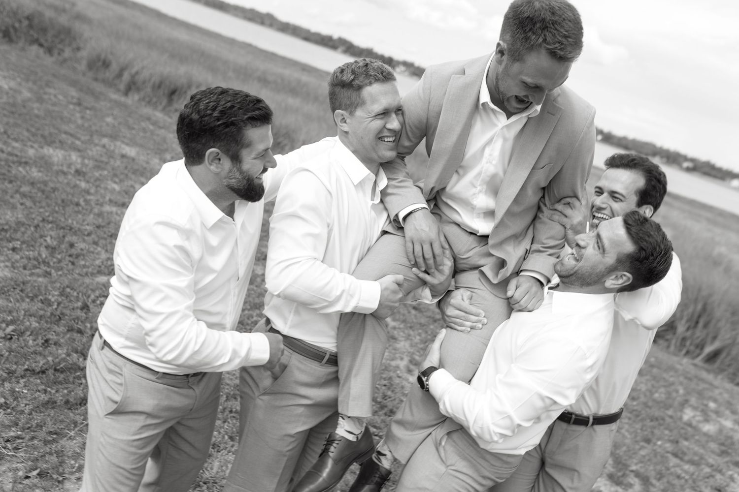 A group in white shirts and khaki pants playfully wrestle and laugh together outdoors on a grassy field.