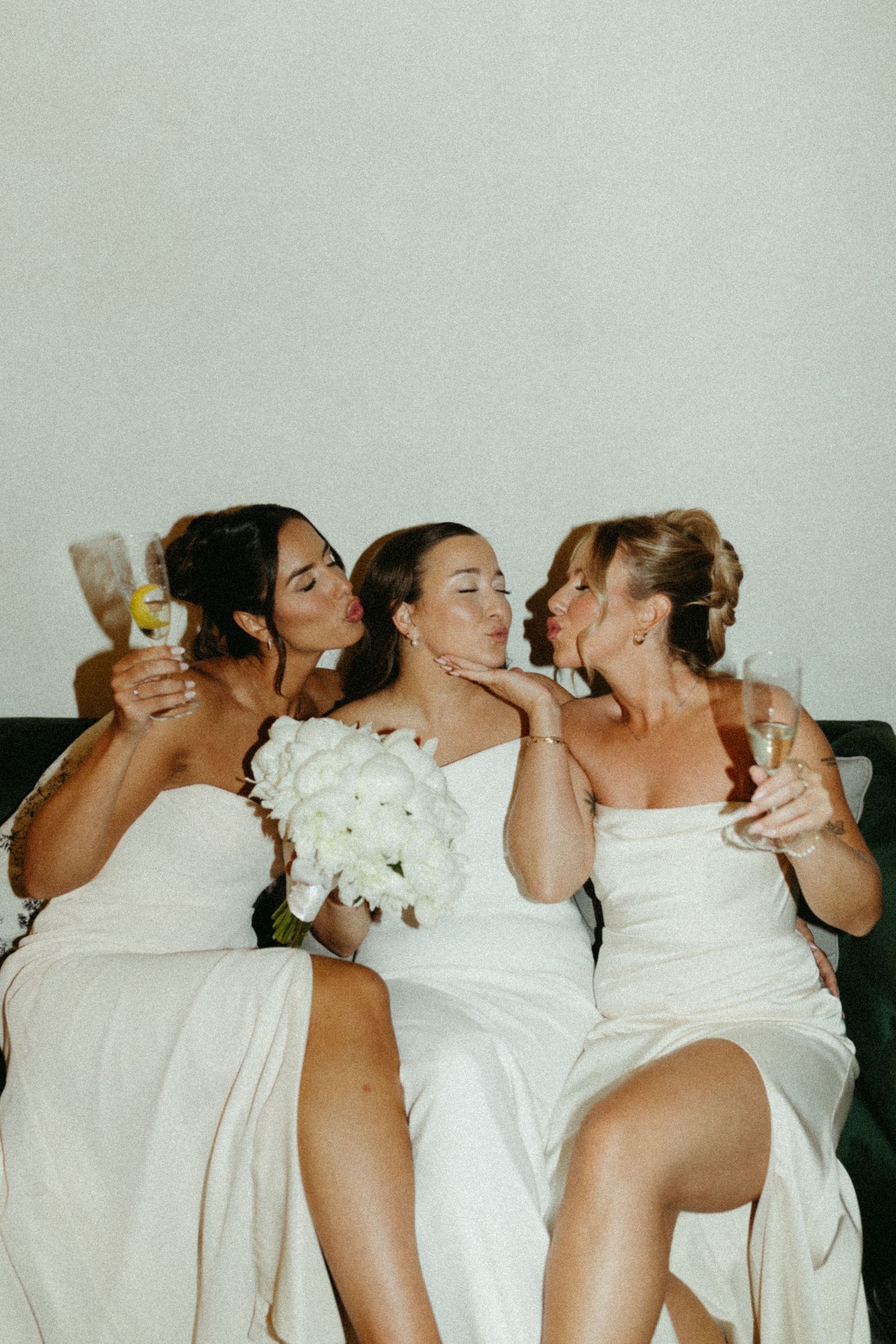 Three women in white dresses sitting together celebrating with drinks and laughter at a wedding celebration.