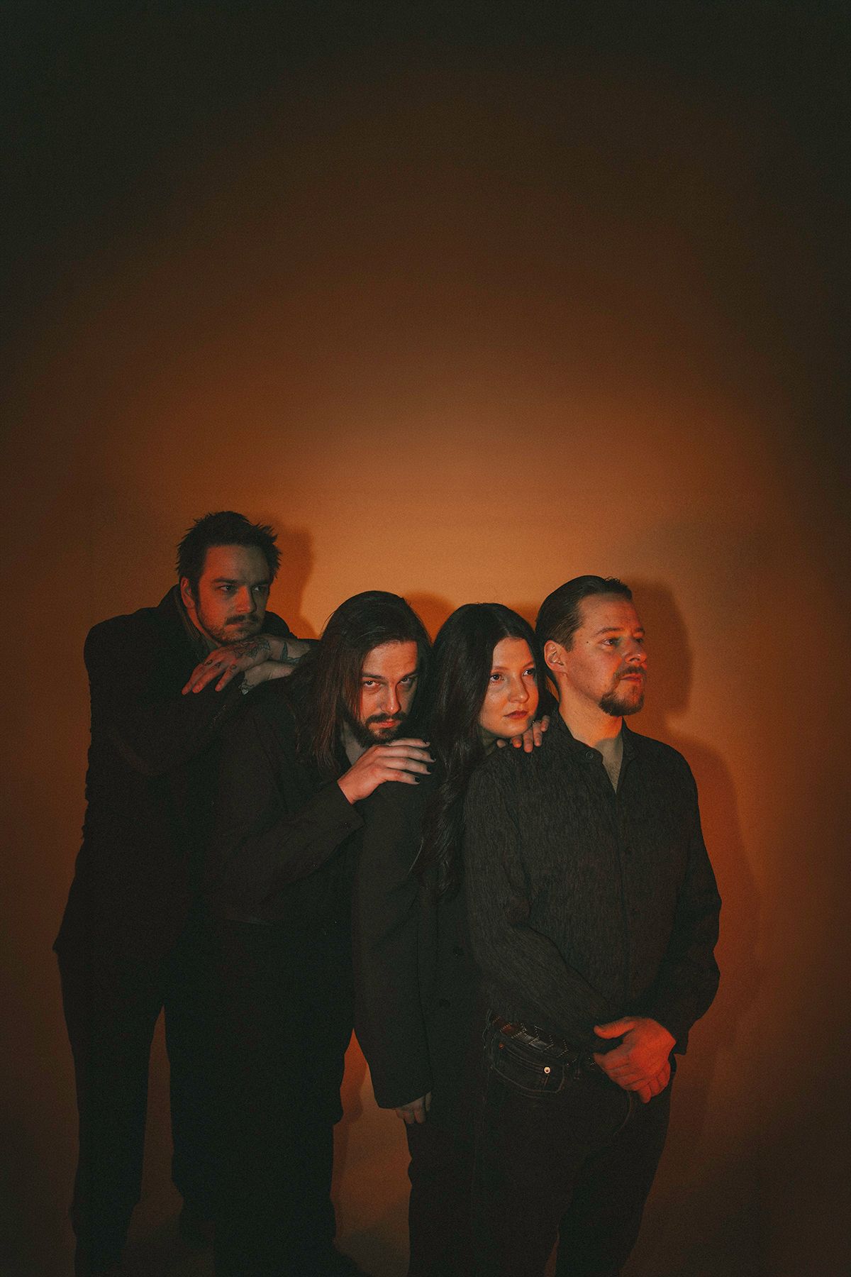 A moody portrait of a four-person music group in black attire against an orange-tinted dark background.