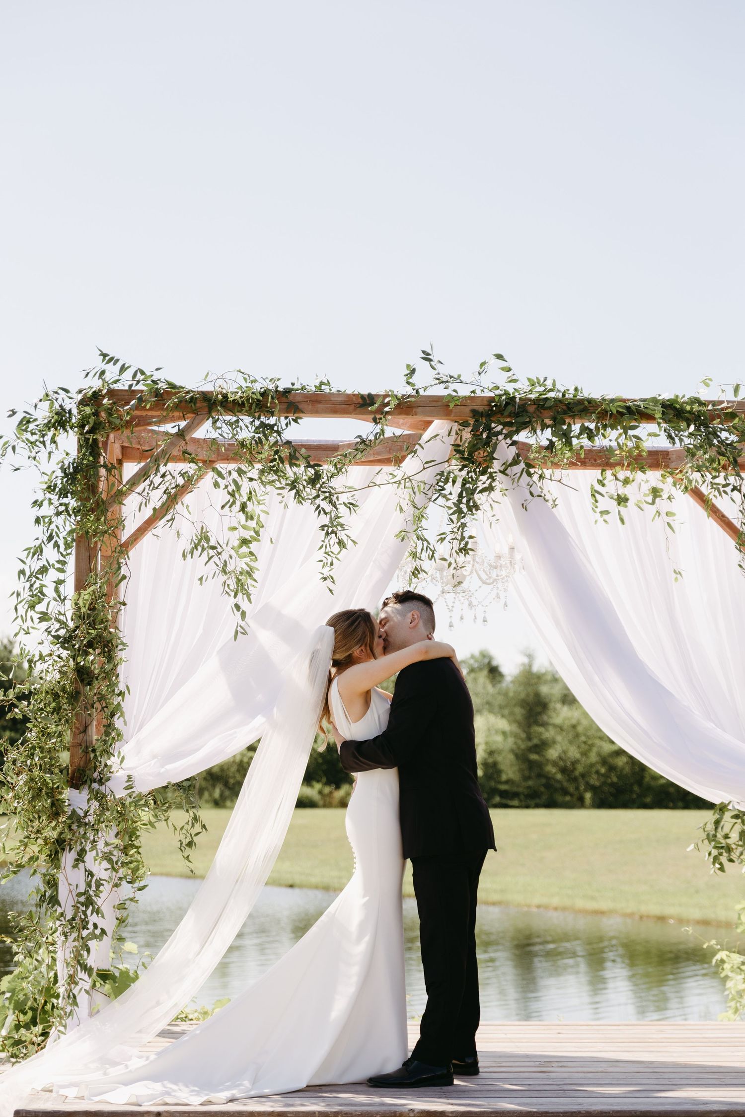 A couple shares a kiss under a white draped wedding arch decorated with greenery beside a pond.