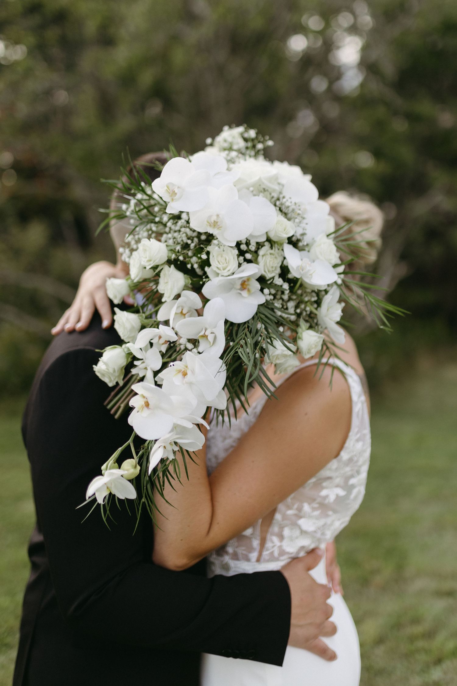 A romantic wedding couple embraces while holding an elegant white floral bouquet with cascading orchids and greenery.