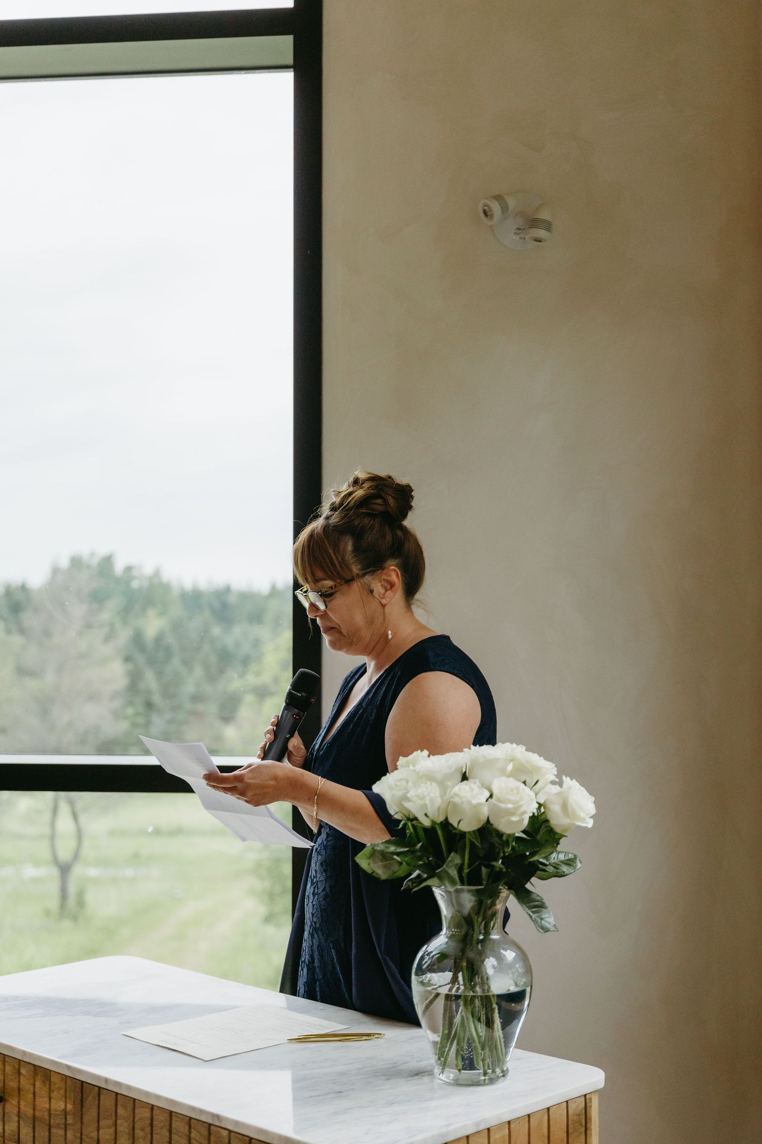 A person in a navy dress stands by a window checking their phone next to a vase of white roses on a counter.