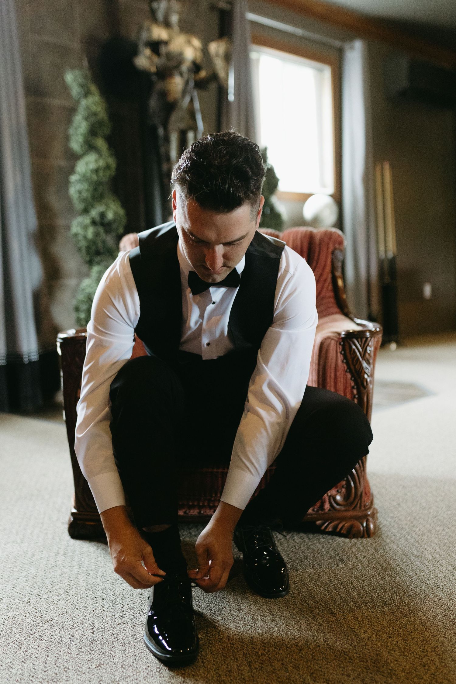 Person in formal attire sits on carpeted floor in contemplative pose near window in indoor setting.