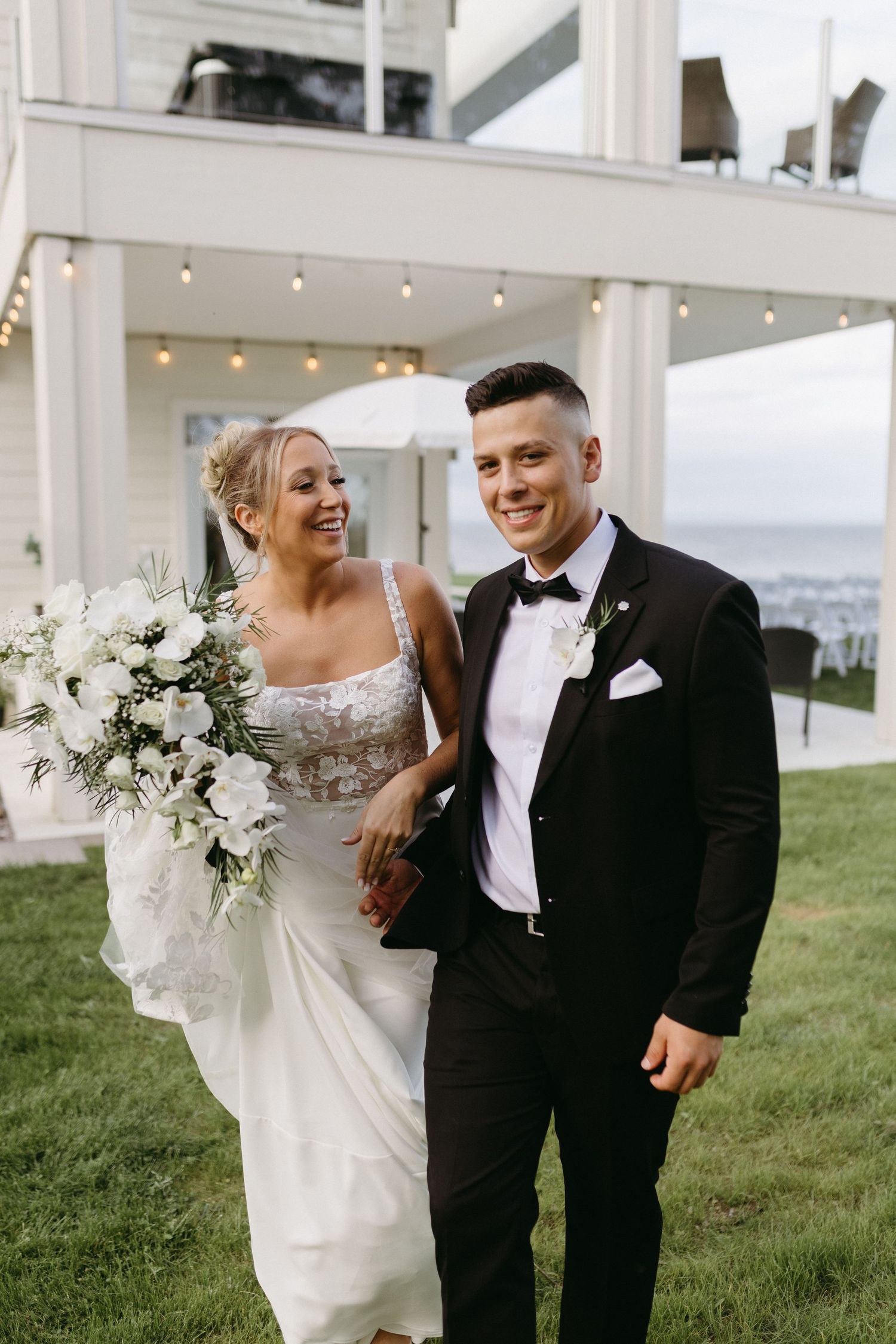 A happy couple shares a joyful moment on their wedding day outdoors with string lights and a white floral bouquet.
