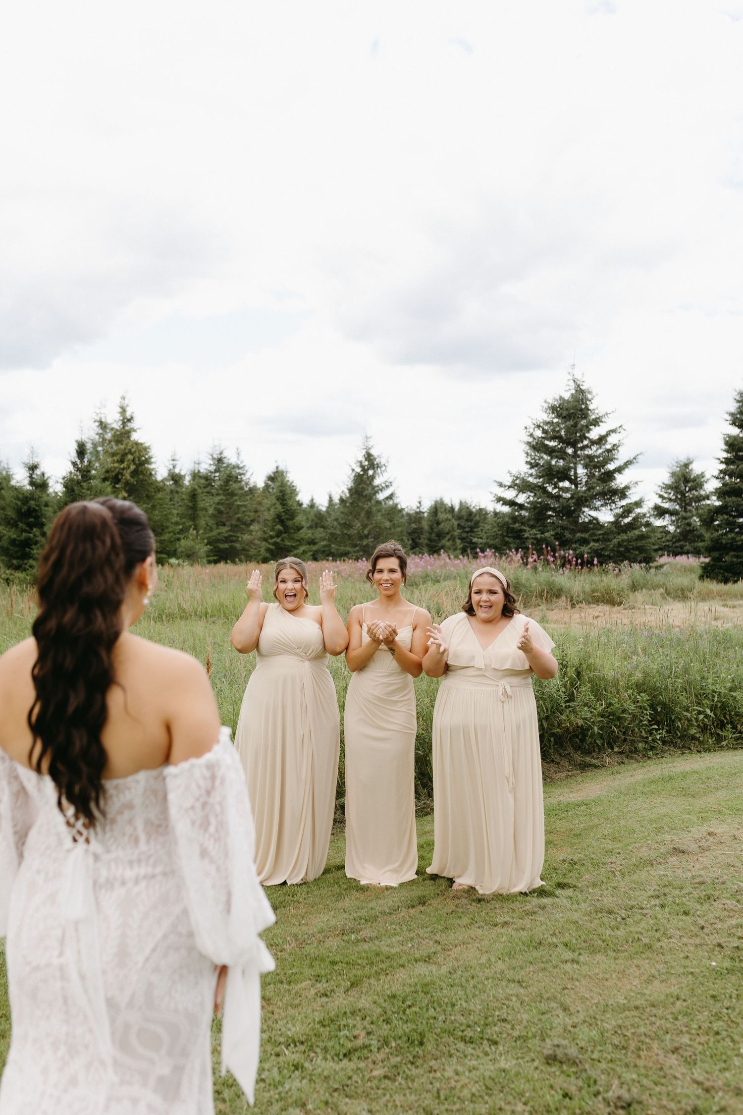 Bridesmaids in champagne-colored dresses share a joyful moment with bride in lace gown on grassy field near evergreen trees.