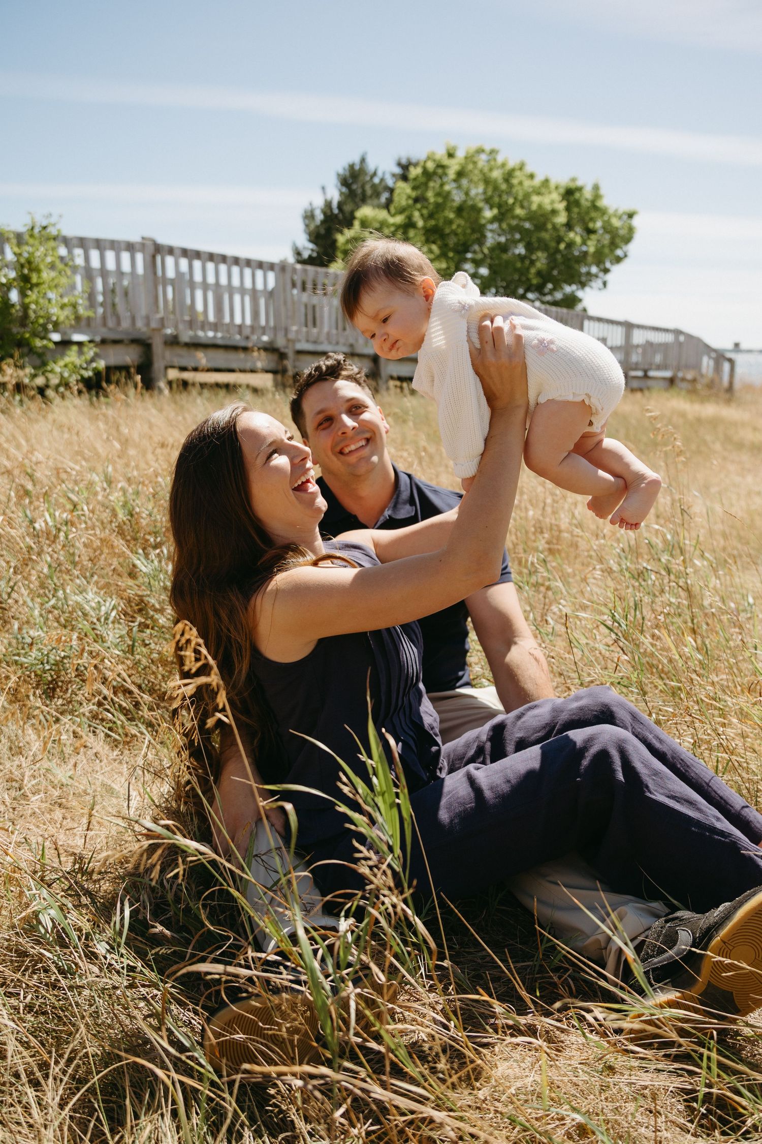 A family relaxes together in tall grass on a sunny summer day with a wooden bridge in the background.