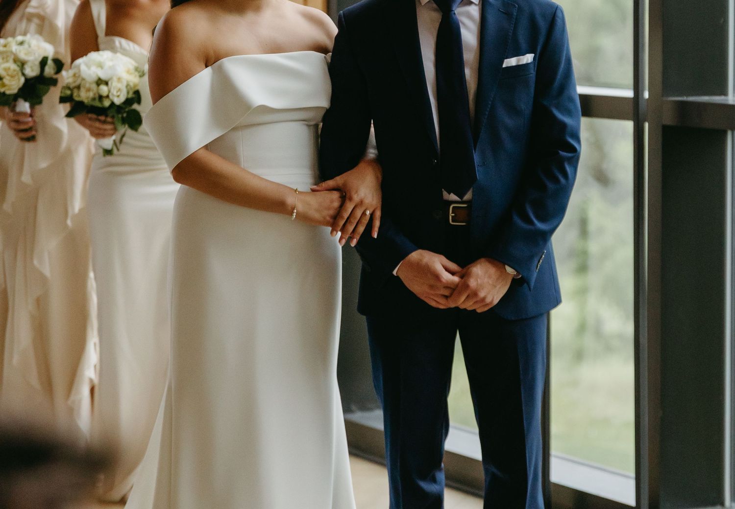 A couple stands together at a wedding, with the bride in white dress and groom in navy suit holding hands by a window.