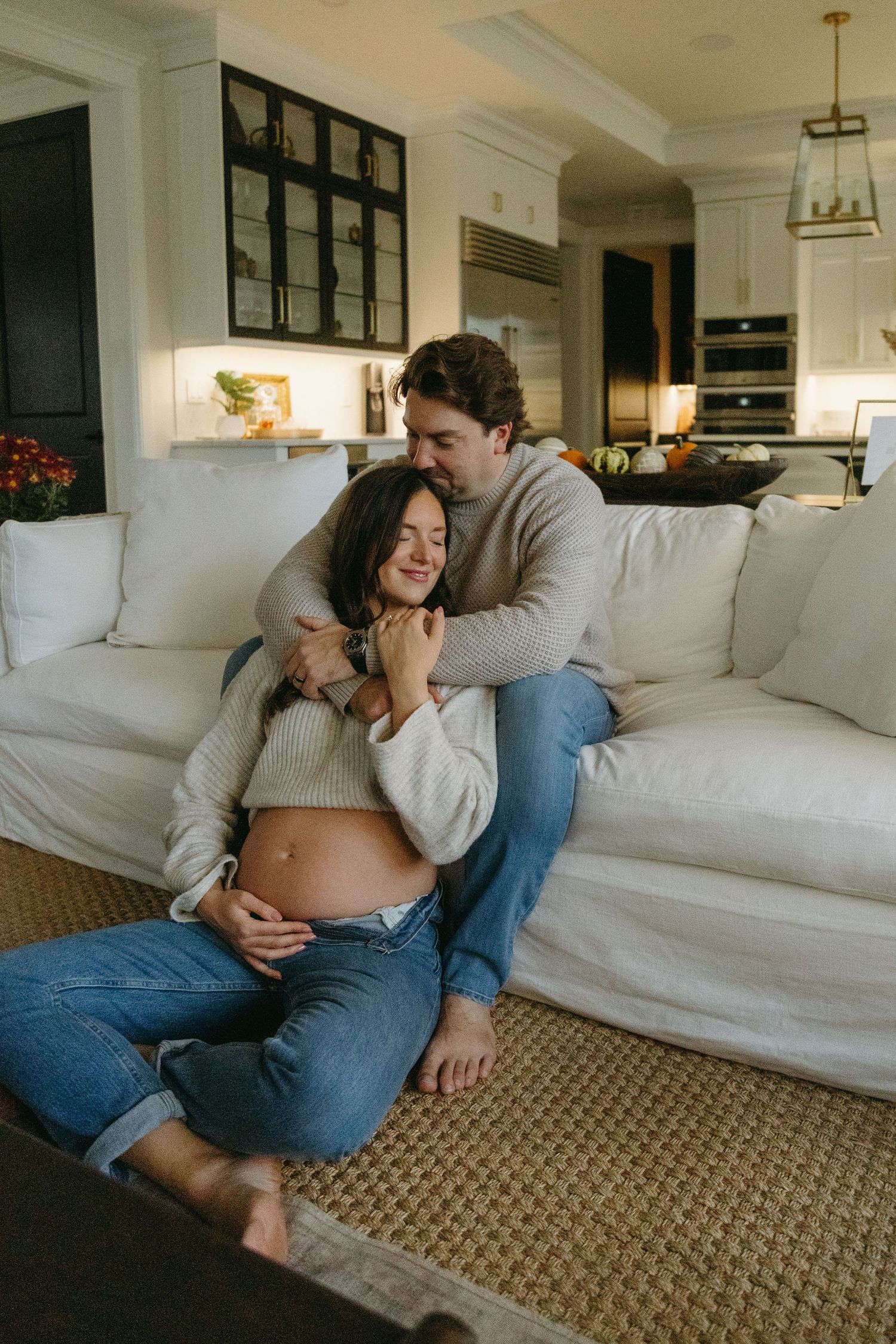 Expectant couple shares intimate moment on white couch in modern kitchen with exposed belly showing pregnancy.