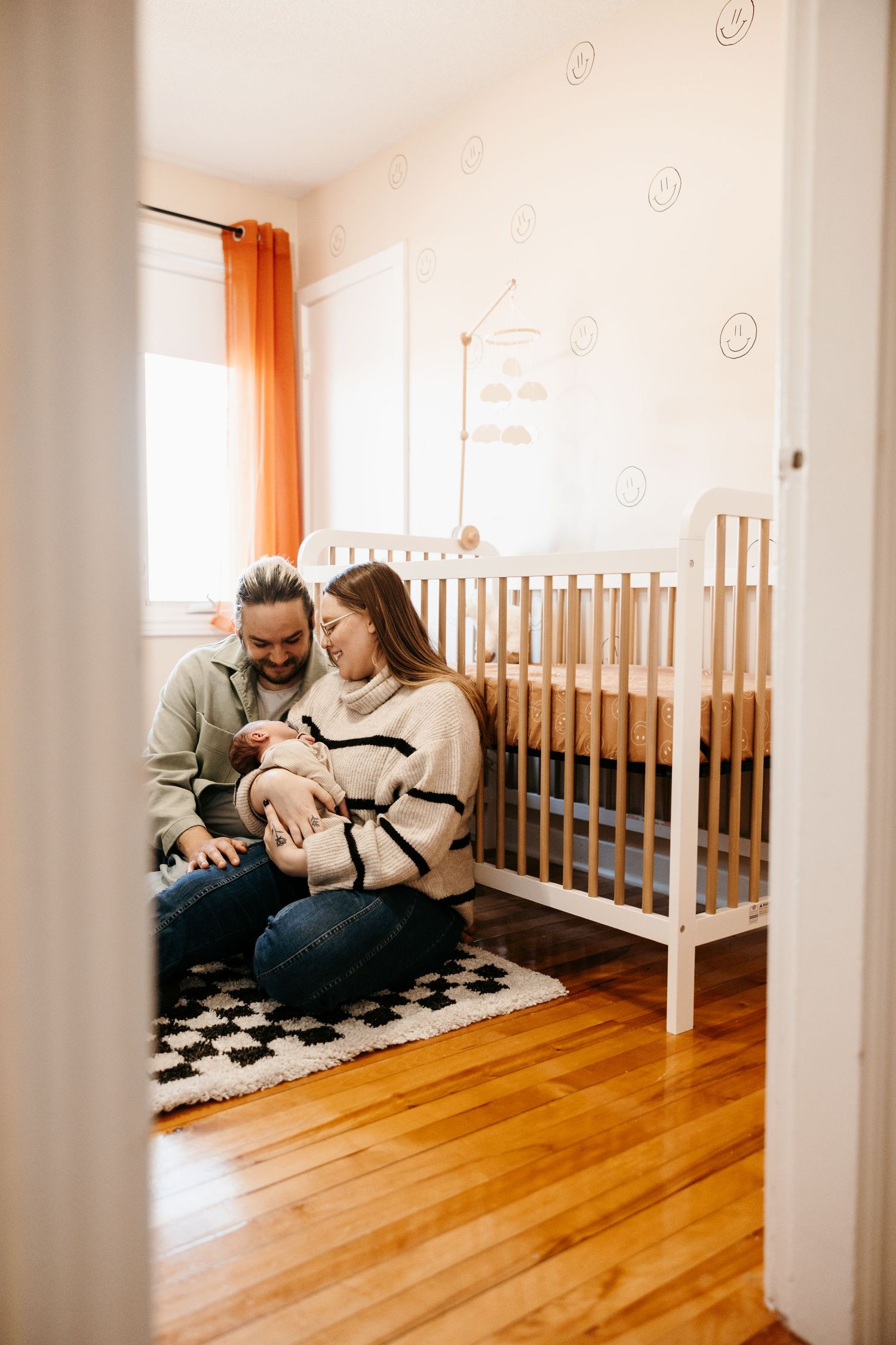 Family relaxing together on hardwood floor in bright nursery room with white crib and orange curtains.