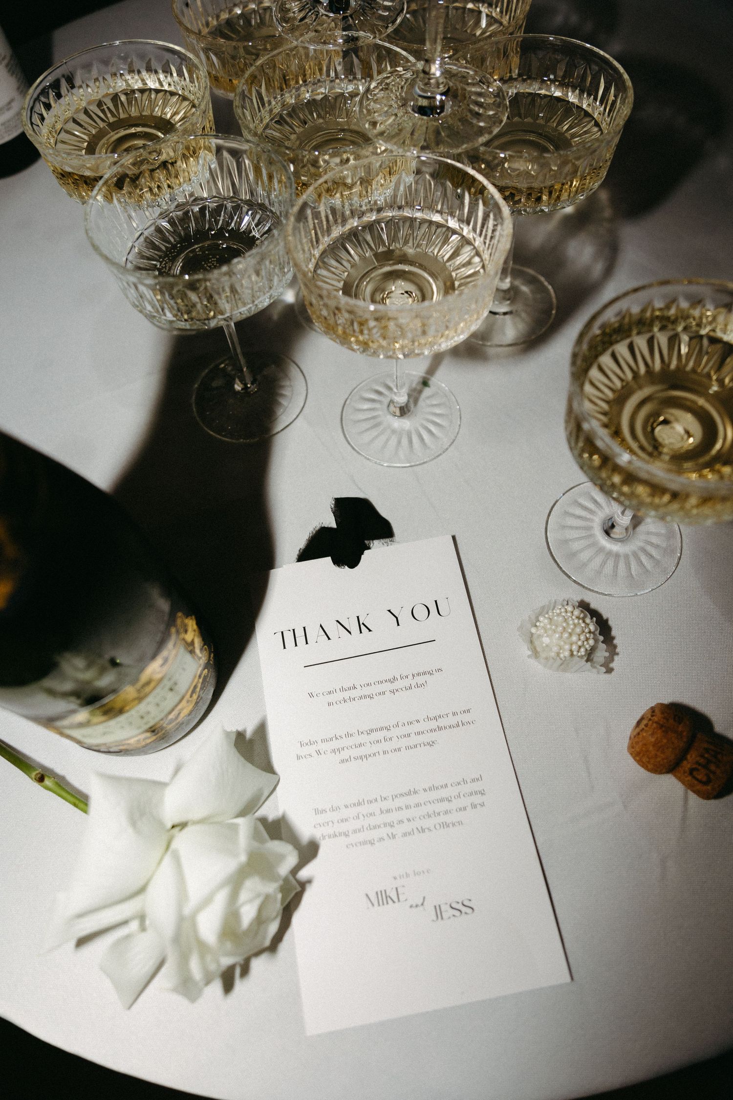 Crystal champagne glasses and elegant thank you cards create an upscale wedding reception setting on a white surface.