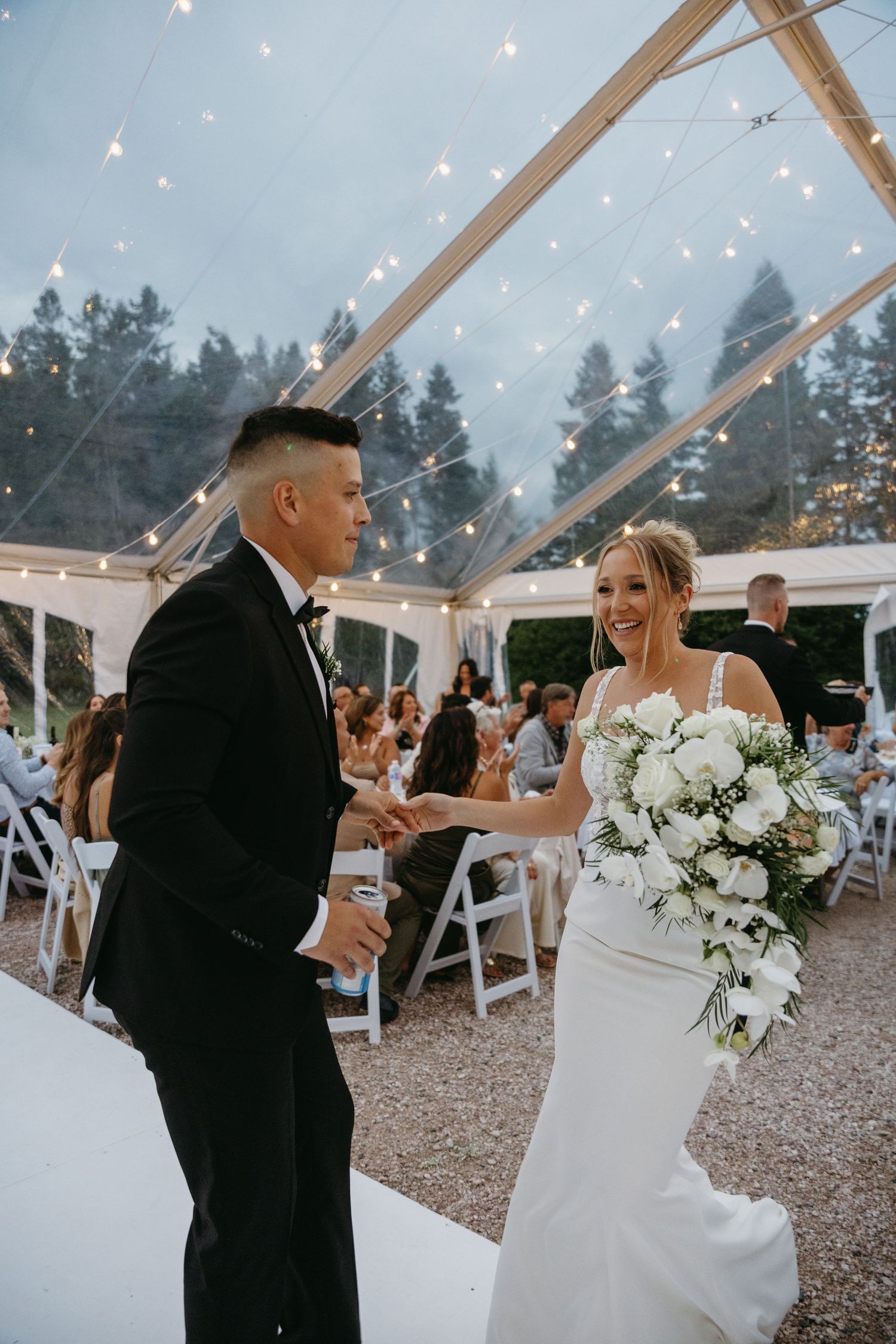 Couple dancing under a clear tent with twinkling lights and white floral decorations at an outdoor wedding reception.