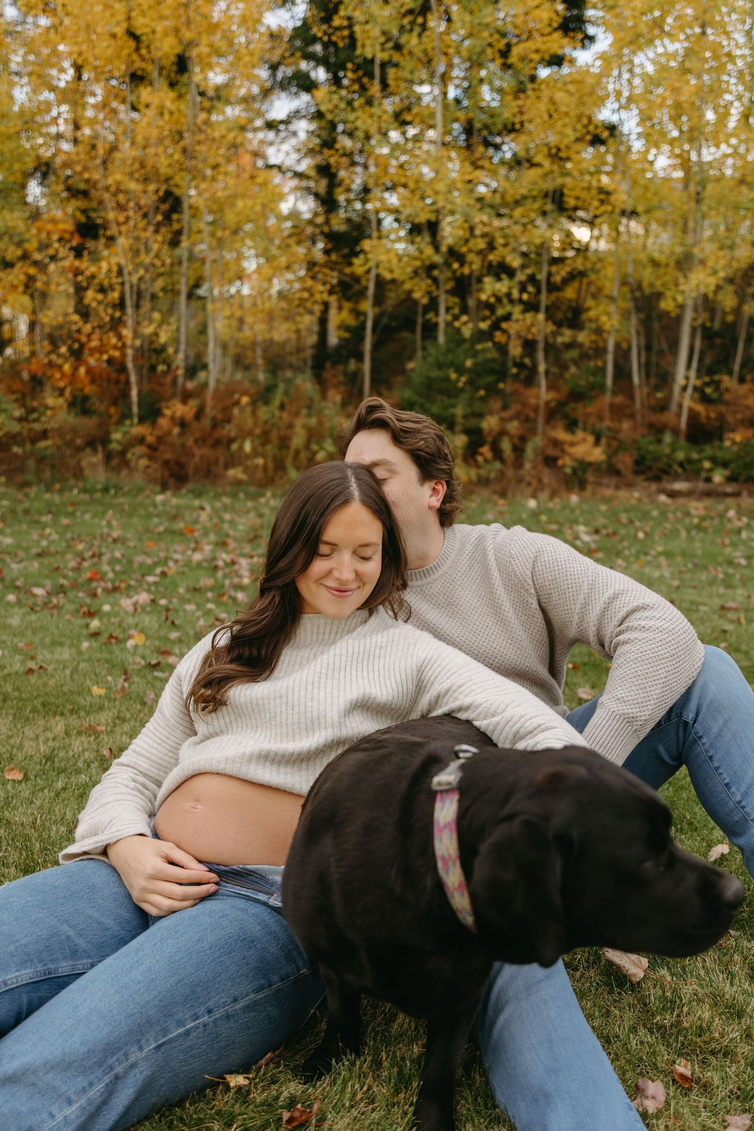 A couple sits on grass with their black dog during autumn, surrounded by golden trees and fallen leaves.