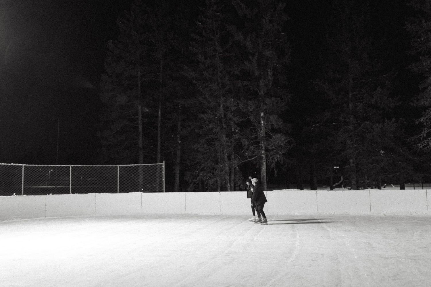 A lone skater glides across a snowy outdoor ice rink at night with dark forest in the background.