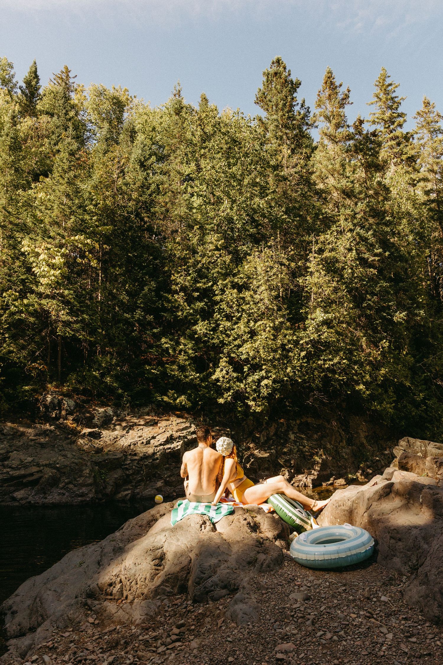 People relaxing on rocky cliffs by a lake with pool floats and evergreen trees in the background during summer.