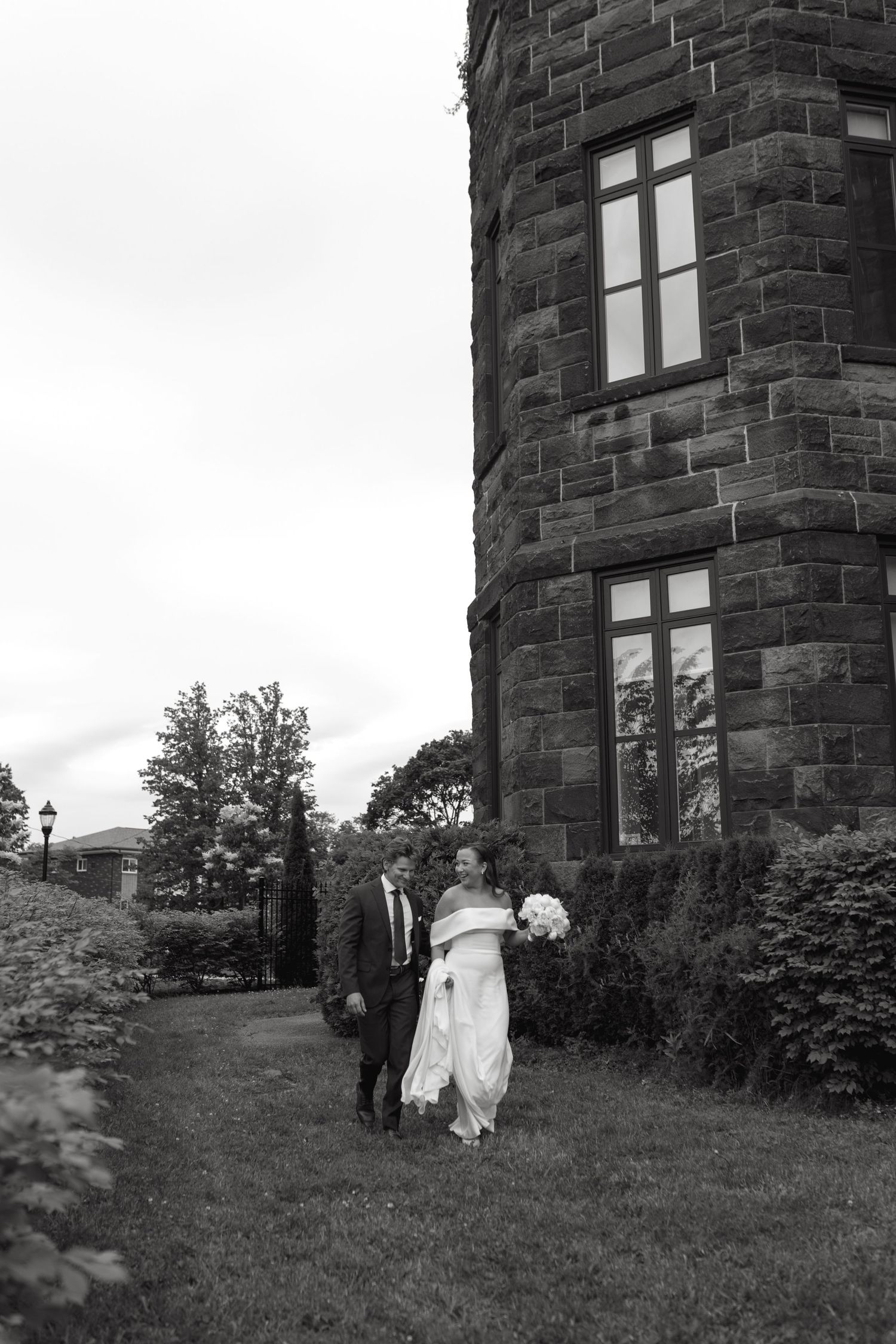 Black and white photo of a couple walking outside a stone castle tower with tall windows on their wedding day.