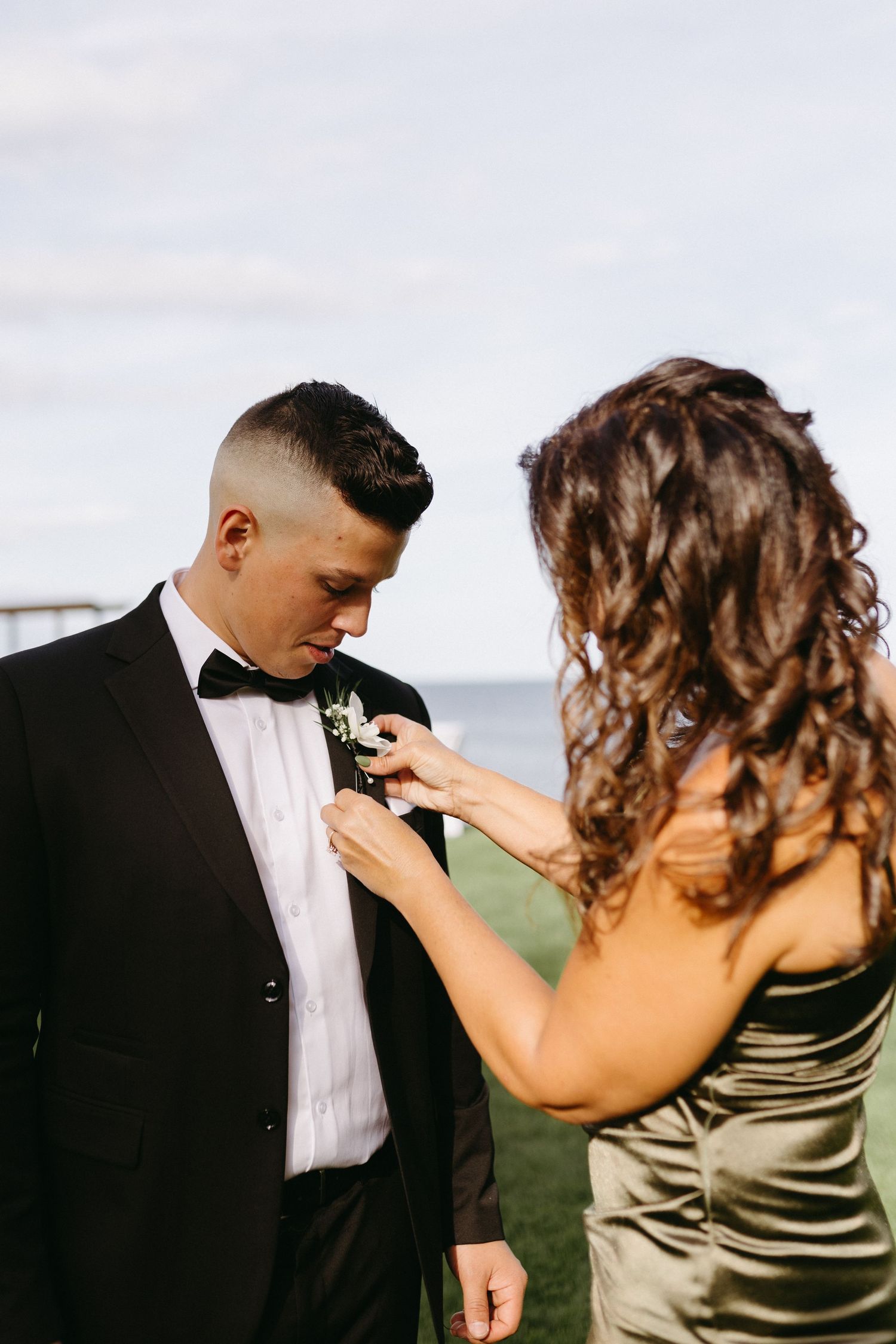 A person in a sage green dress pins a white boutonniere onto someone wearing a black tuxedo at an outdoor event.