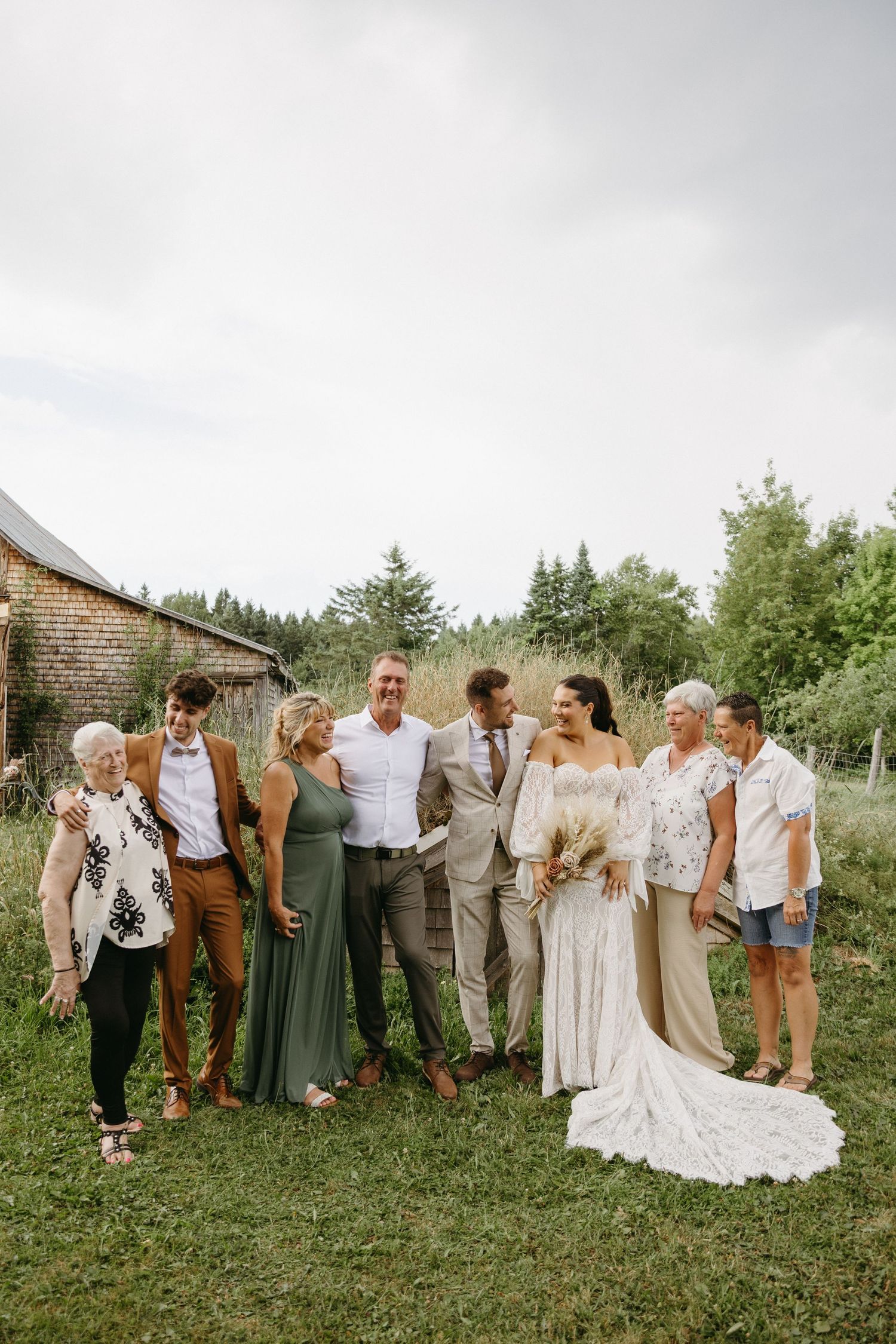 Wedding party poses together on a grassy hillside wearing earth tones and white dresses for outdoor summer celebration.