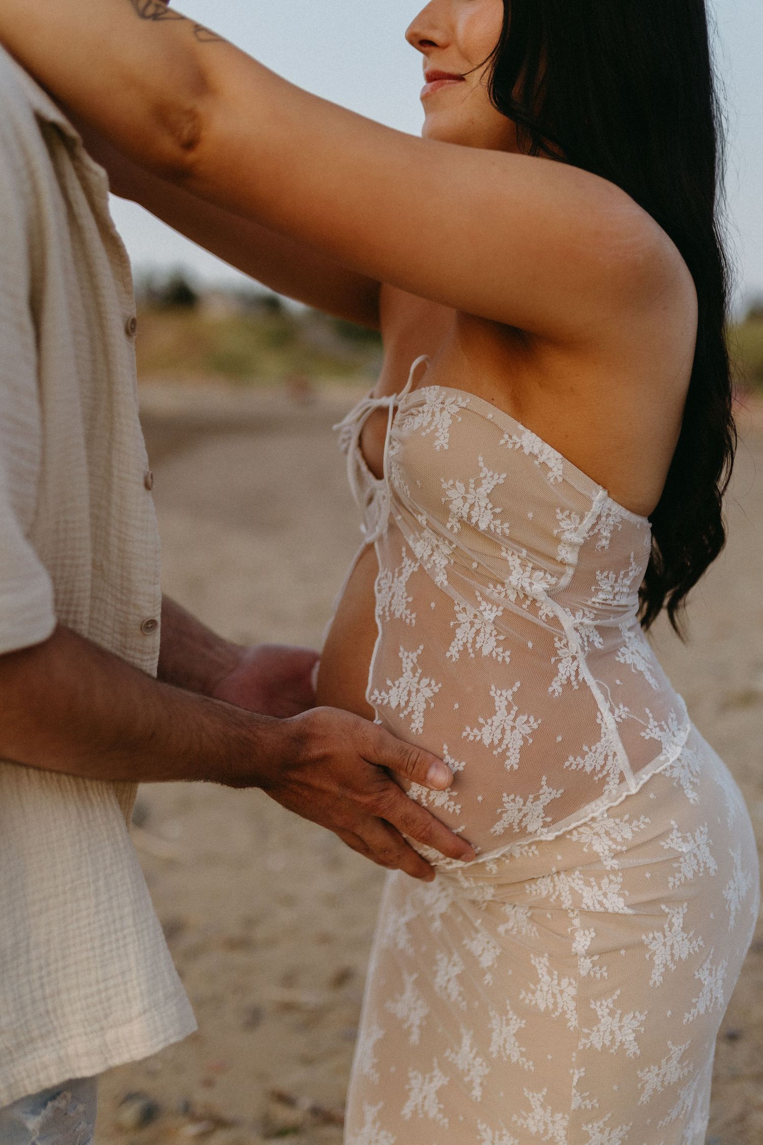 A close-up shot of a couple embracing, with focus on a delicate white lace dress against a beach backdrop.