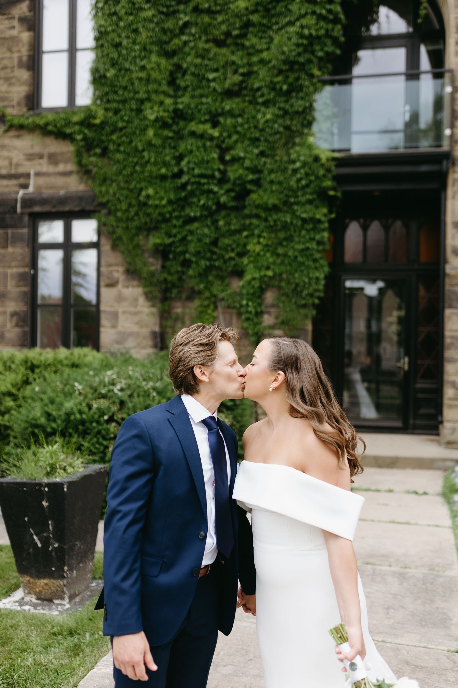 A couple in wedding attire share a romantic kiss outside a stone building with ivy-covered walls.