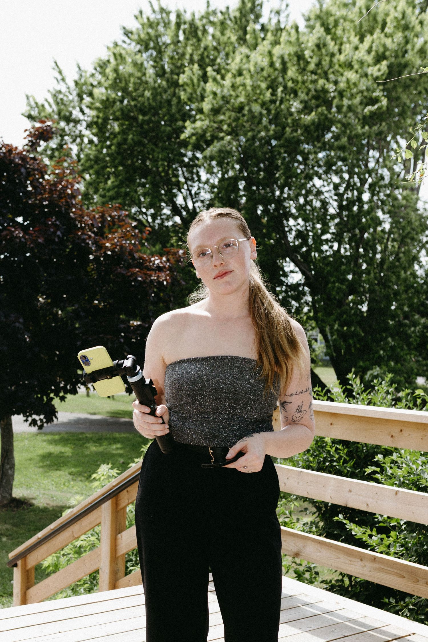 A photographer in a black strapless top and pants stands on a wooden deck surrounded by greenery.