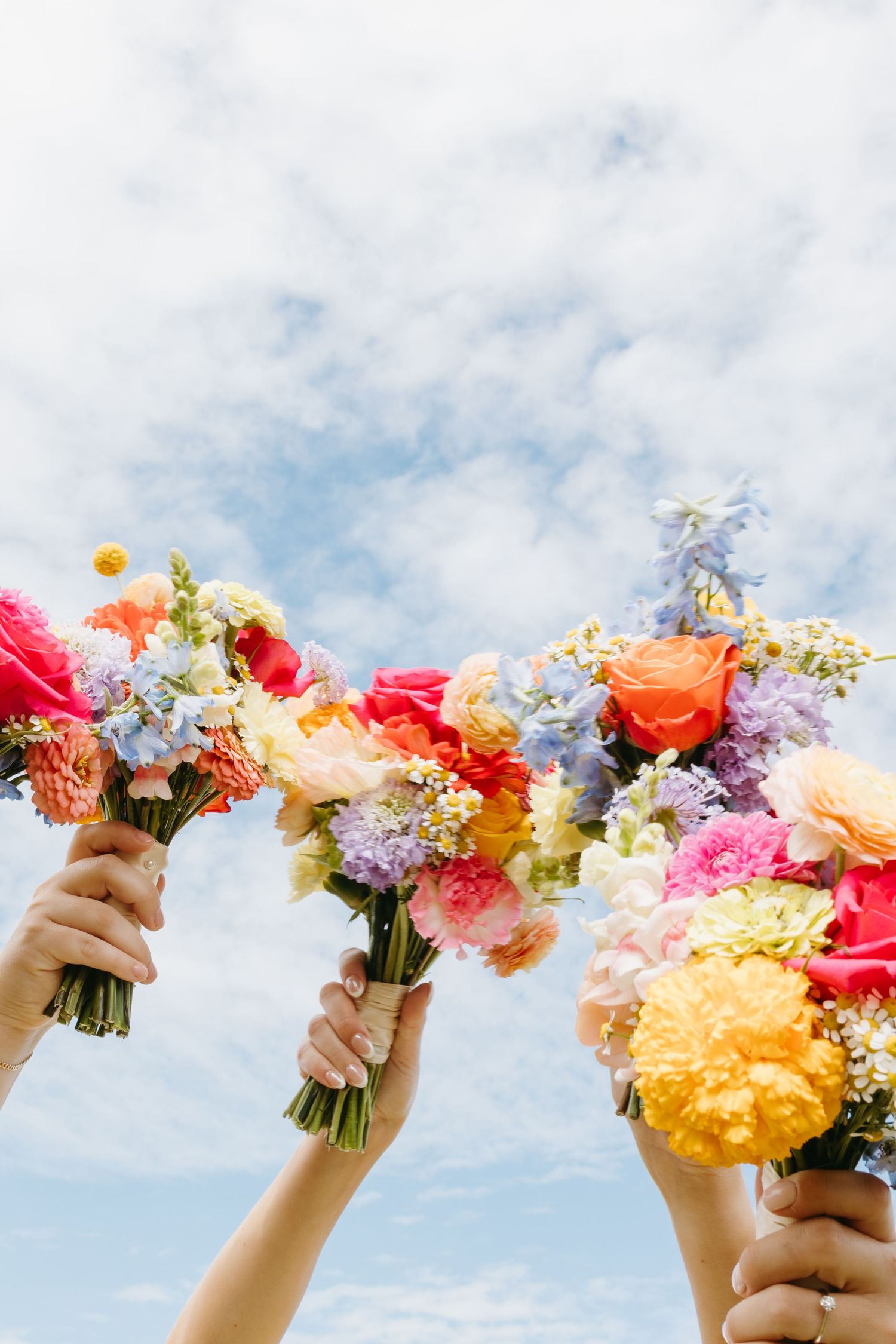 Three hands hold up colorful bouquets of mixed flowers against a bright blue sky with white clouds.