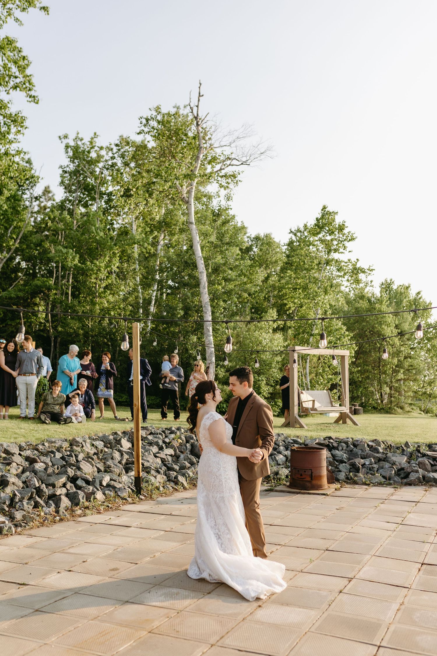 A couple shares their first dance on a stone patio surrounded by trees and guests at an outdoor wedding venue.