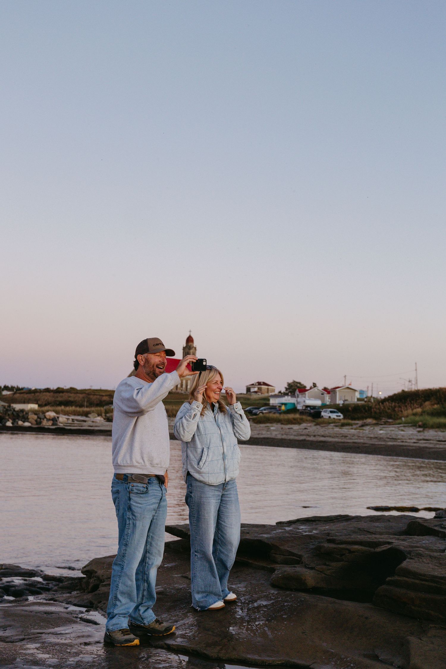 Two people in denim outfits stand together on a rocky beach at sunset with boats and houses visible in the background.