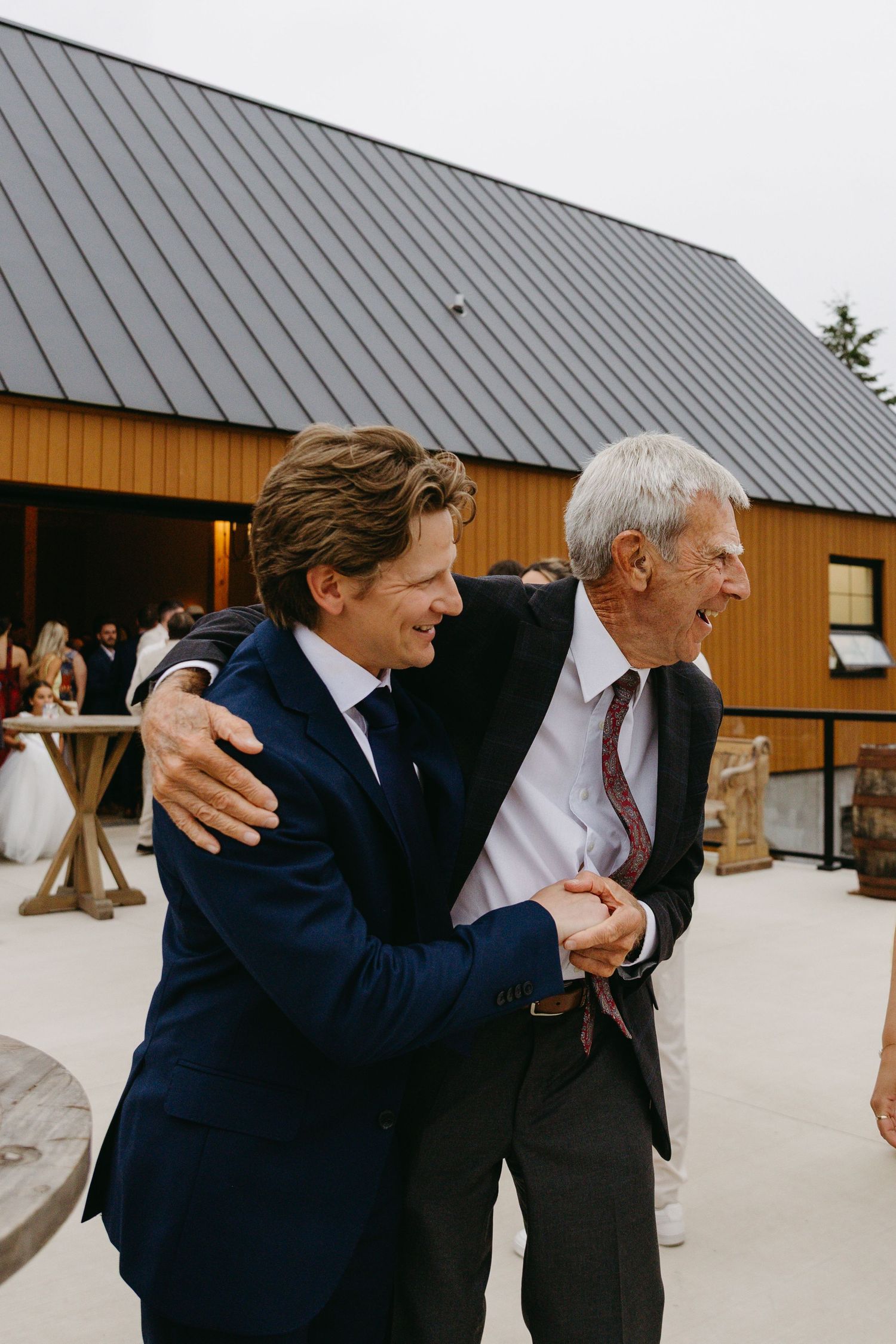 A sequence of candid moments showing two men in suits embracing warmly outside a wooden barn building.